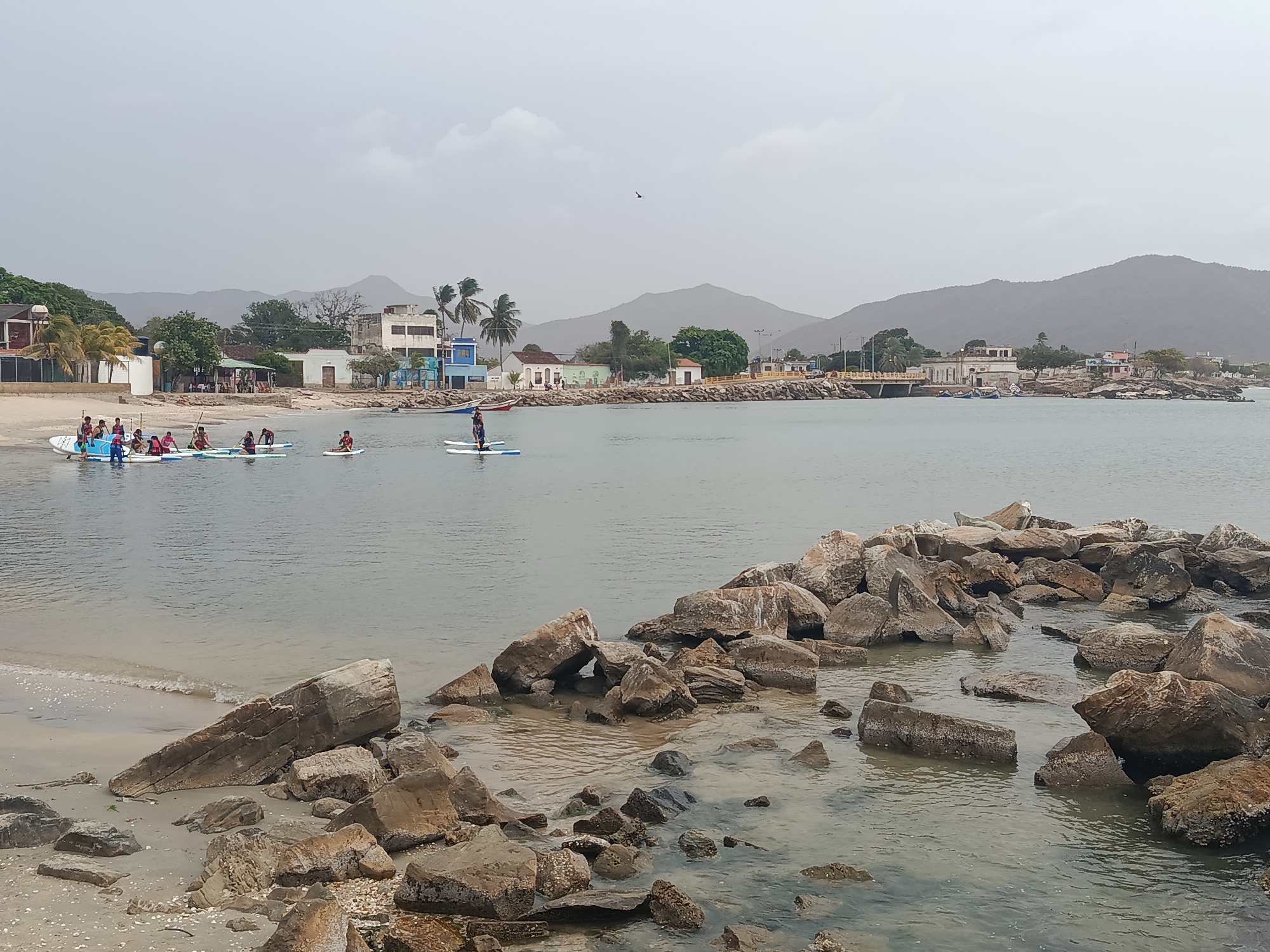 The image depicts a coastal scene in Juan Griego, Venezuela, characterized by a hazy sky and calm waters. In the foreground, a rocky barrier extends into the water. To the left, a sandy beach is visible, and several people are participating in paddleboarding activities. One group is clustered on a large paddleboard near the shore, while another individual stands on a separate paddleboard closer to the center of the frame. The background shows a line of buildings along the shoreline, interspersed with trees, and a range of hills in the distance.