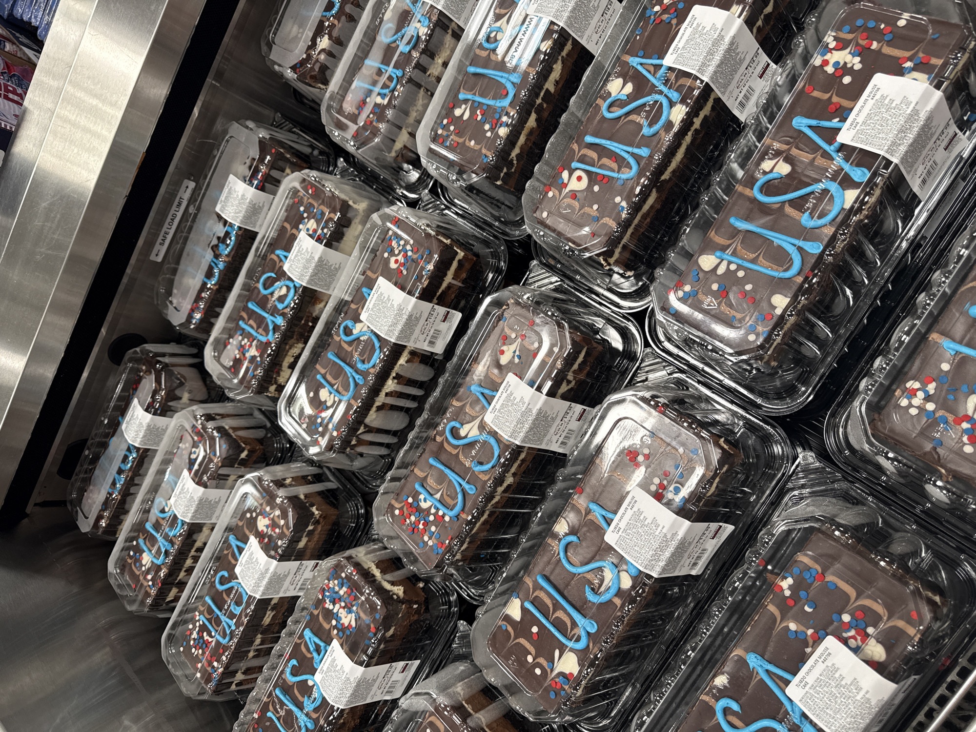 The image shows a close-up, overhead view of several individually packaged chocolate mousse cakes on display, likely in a retail environment such as a grocery store or bakery. The cakes are rectangular and are decorated with blue icing that spells out "USA" on each. They are also adorned with red, white, and blue sprinkles, and swirled chocolate and white icing. The cakes are housed in clear plastic containers with white labels indicating nutritional information and product details. The background is dominated by the reflective surface of a stainless steel display case and hints of other products on shelves above. The setting appears to be the City of Yonkers, United States of America, suggesting a patriotic or holiday-themed display. There are no people visible in the image.