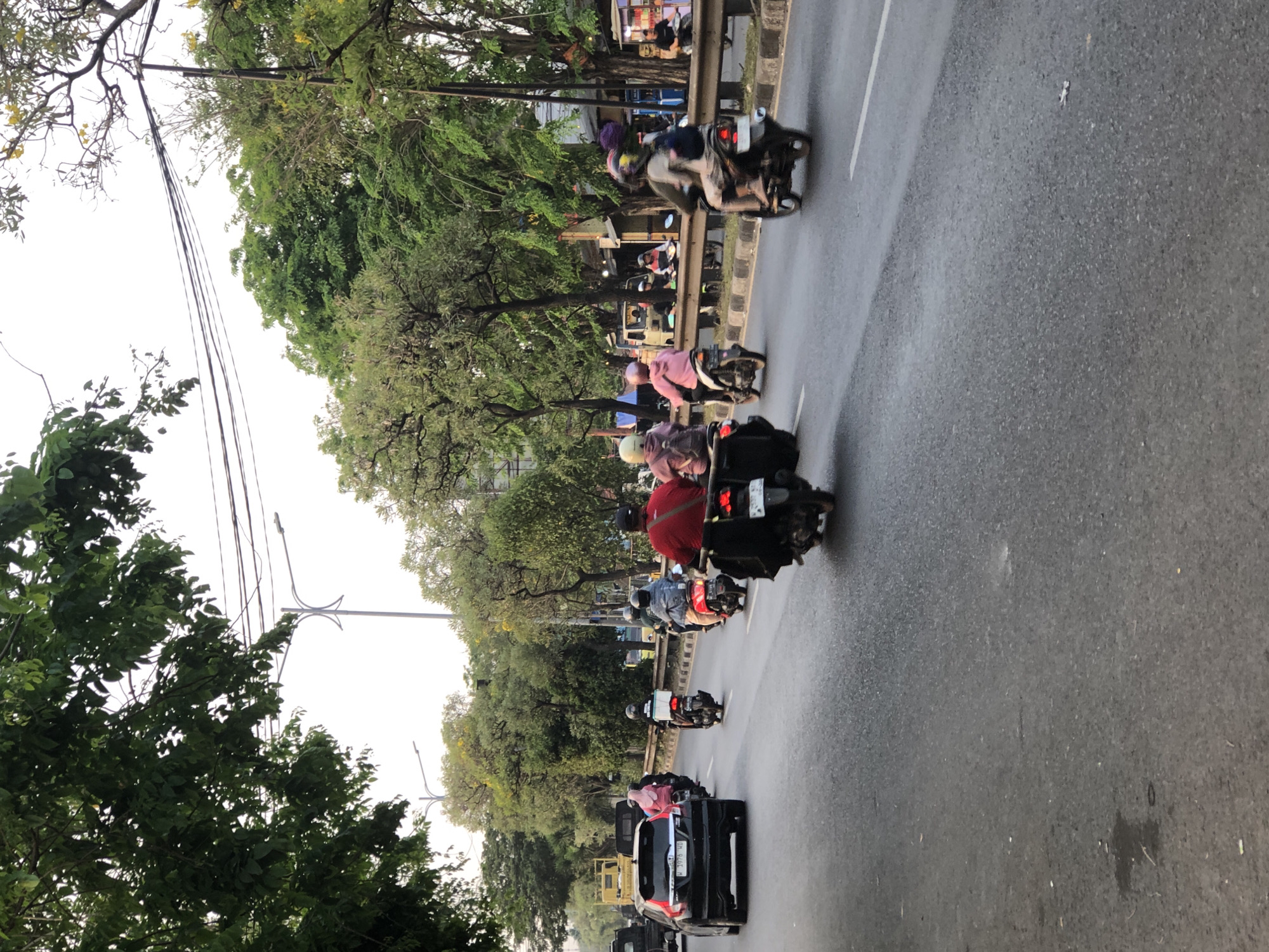 The image depicts a street scene in Kludan, Indonesia, characterized by moderate traffic. In the foreground, the asphalt road shows signs of wear, with some minor stains. Several motorcycles are in motion, with riders wearing helmets and some carrying passengers or cargo. A black car is visible on the left side of the road, and further down, a yellow truck is parked. The surrounding environment features lush green trees lining the street, with some yellow flowers blooming. Overhead, utility wires and streetlights are present. The sky is overcast.