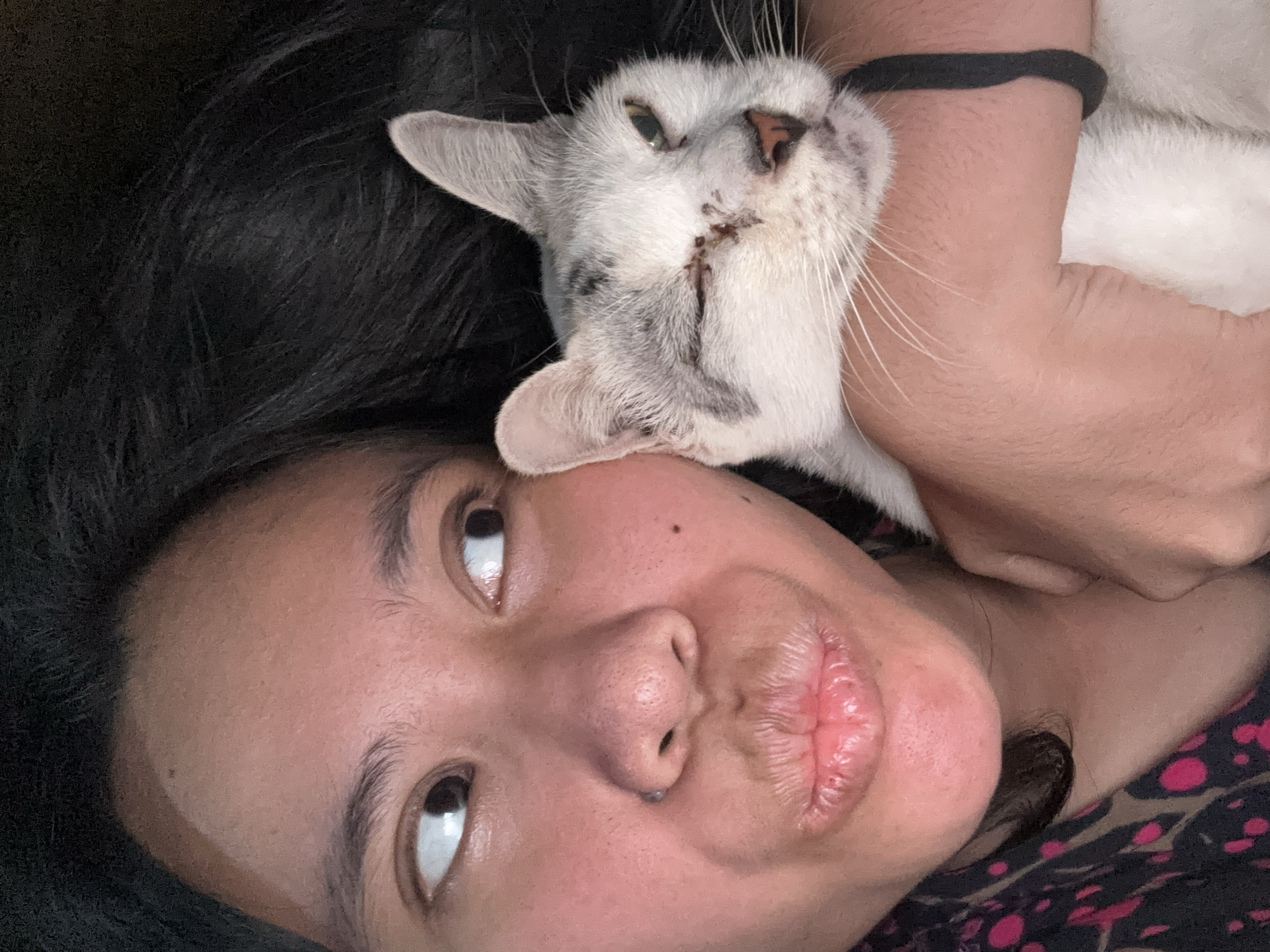 A close-up selfie shows a person with dark hair and dark eyes, puckering their lips and looking to the left. They are holding a white and grey cat, which is also looking to the left and appears to have an injured eye. The background is dark and indistinct, suggesting an indoor setting. The person is wearing a black shirt with a pink pattern. The image was taken in East Jakarta, Indonesia.