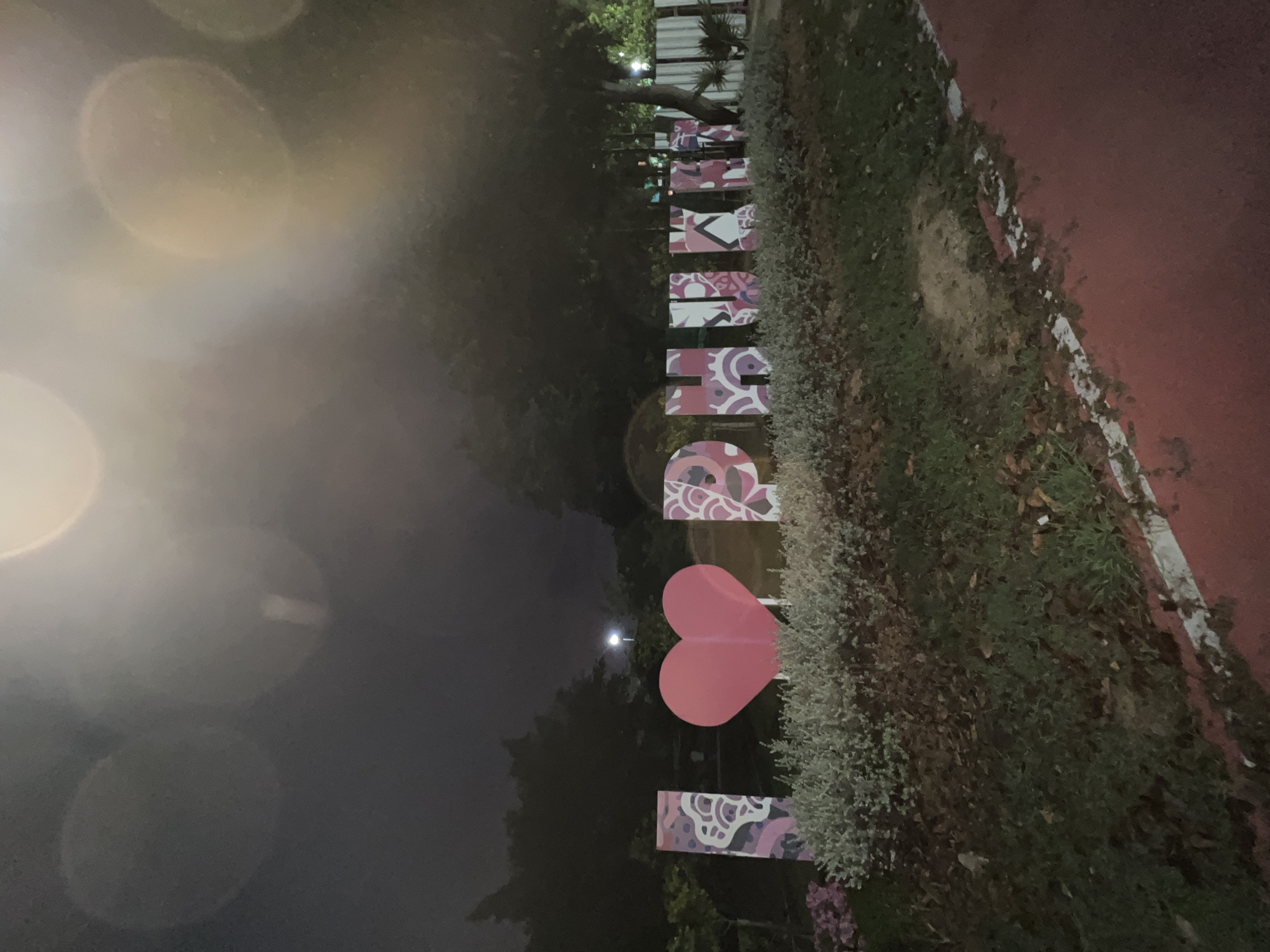 The image shows a nighttime view of a sign spelling out "PHUKET" in Phuket City Municipality, Thailand. The letters are decorated with a pink and white design, with the letter "H" replaced by a pink heart shape. The sign is positioned near a walkway with a red surface and is surrounded by vegetation. A light source is visible in the background. The foreground of the image is blurred, possibly due to lens flare or lighting conditions.