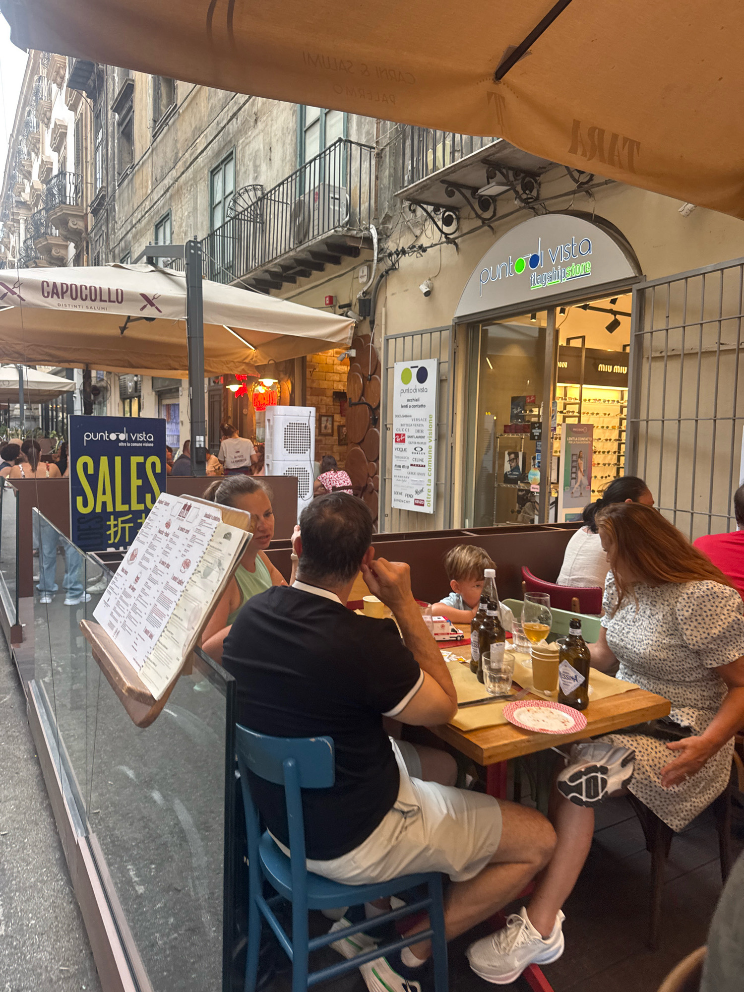 The image depicts an outdoor dining area in Palermo, Italy, likely on a street or pedestrian walkway. In the foreground, a group of people are seated at a wooden table. On the table are several bottles of beer, glasses, and plates with food remnants. A young boy is seated at the table, and an adult woman with red hair is to his right. A man wearing a black shirt and white shorts is seated opposite them, and his back is mostly to the camera. A menu is propped up on an easel in front of the man. In the background, a building with apartments and balconies lines the street. A storefront with the sign "punto di vista flagshipstore" is visible. Adjacent to the storefront, a blue sign with yellow lettering reads "SALES" and some Japanese characters. Umbrellas provide shade over the dining area. The overall atmosphere suggests a casual dining experience on a warm day.