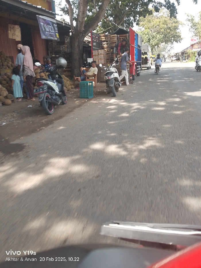The image captures a bustling roadside scene in Patrol, Indonesia, on a bright, sunny day, dated February 20, 2025, at 10:22 AM. The perspective is from a moving vehicle, with a portion of its dashboard or mirror visible in the foreground.
On the left, a street stall is set up, overflowing with a large pile of durians, indicating a fruit market or vendor. Several people are present at the stall: a man in a blue shirt, a woman wearing a hijab and a long dress, and a small child in a light blue dress standing beside her. Another individual is partially visible further inside the stall. A black and red motorcycle is parked in front of the durian display.
In the midground, a large tree provides shade over the roadside. Underneath it, a man in a light-colored shirt is seated on a wooden chair, resting, with a green plastic crate nearby. Adjacent to him is a parked blue and red truck, heavily loaded with wooden crates, suggesting it's used for transporting goods, possibly the durians. A person is seen standing near the back of the truck, possibly assisting with loading or unloading. Another white scooter is parked closer to the truck.
The road itself is asphalt, marked with dappled shadows from the overhead trees. In the background, the road stretches into the distance with other vehicles, primarily motorcycles, and more buildings and trees lining the street. The sky appears clear and bright, consistent with the strong shadows.
Visible text includes a partially obscured sign on the stall that reads "MATI PA" and a banner with "T'WIZ'Z" along with smaller text. A watermark at the bottom left indicates "vivo Y19s Akhtar & Arvan 20 Feb 2025, 10.22." The overall atmosphere suggests a typical busy market day in a tropical or subtropical region.