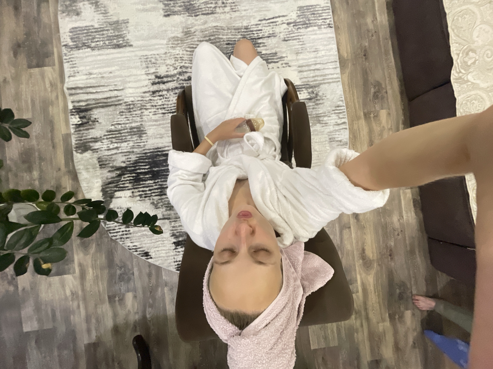 The image shows a woman in a white bathrobe and a pink towel wrapped around her head, sitting in a brown chair. She has her eyes closed and her lips are slightly pursed. She is holding a small object, possibly a cosmetic product, in her right hand. A patterned rug is visible behind her, and a plant hangs from above. The floor is made of wooden planks. The image appears to be taken indoors, in what could be a home or a spa. There is no discernible indication of the city of Sharkan, Russia.