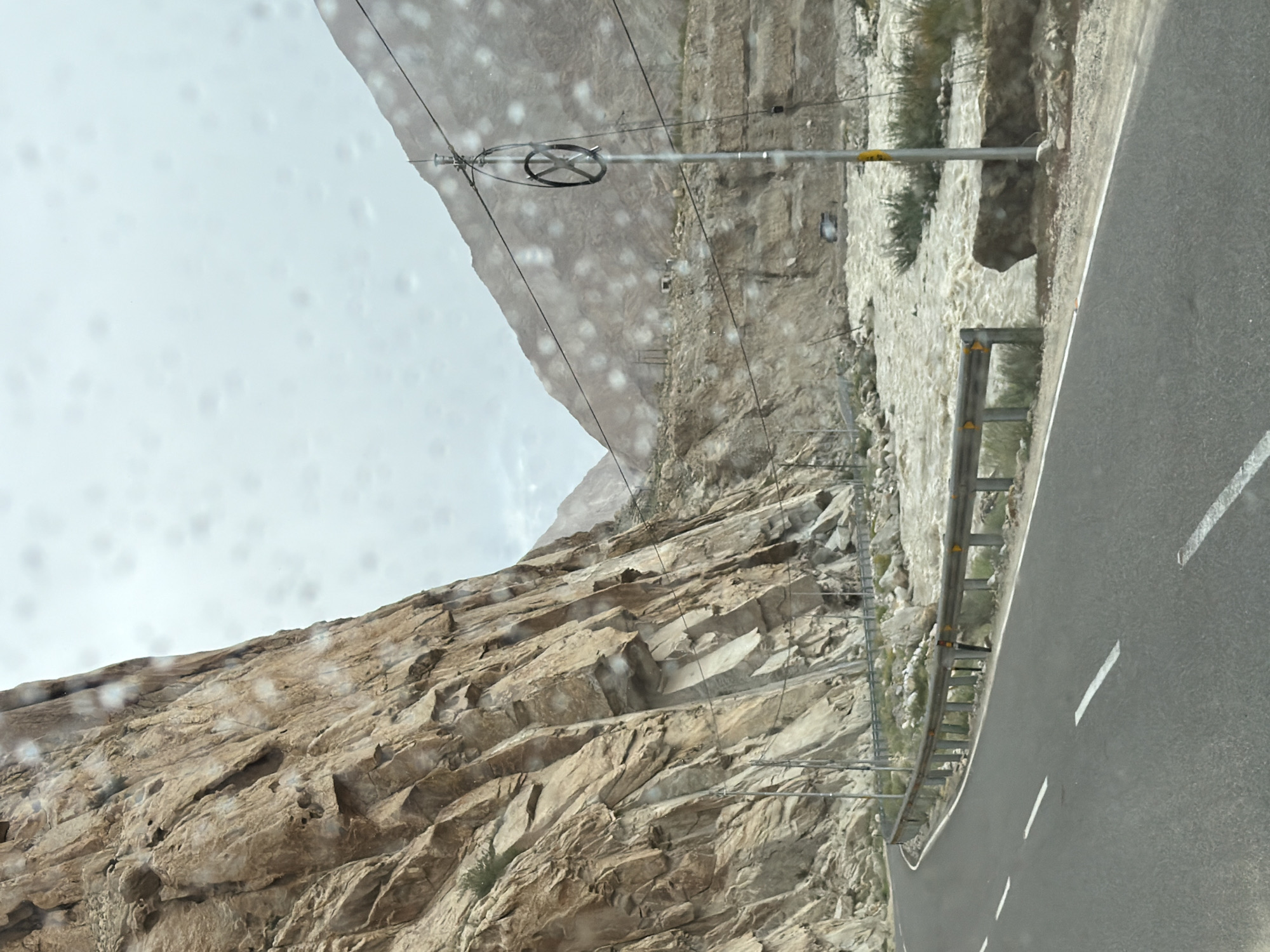 The image shows a winding asphalt road with white lane markings, curving alongside a steep, rocky cliff face. A metal guardrail runs along the edge of the road, separating it from a fast-flowing, muddy river. In the background, more mountains are visible under a cloudy sky. Utility poles with wires are present along the roadside, one with a circular device attached. The presence of raindrops on the lens suggests the photo was taken during wet weather. There are no people visible in the image. The scene appears to be a remote, mountainous area.