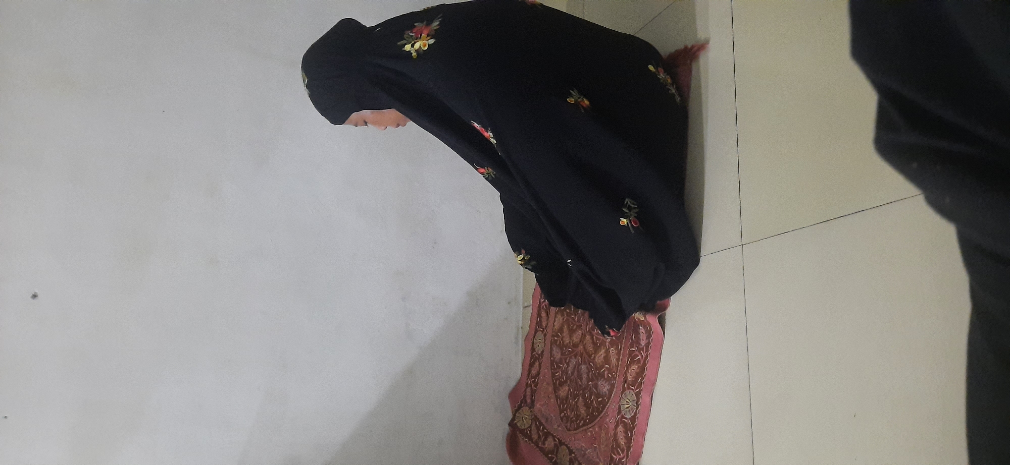 In an indoor setting, a person wearing a black hijab with embroidered floral patterns is seated on a prayer mat. The mat is red with intricate gold and brown designs. The person is facing to the left of the frame, with their head bowed. The background consists of a plain, off-white wall and a light-colored tiled floor. The lighting suggests an indoor environment, possibly during the day or with artificial light. The location is identified as Cisauk, Indonesia.