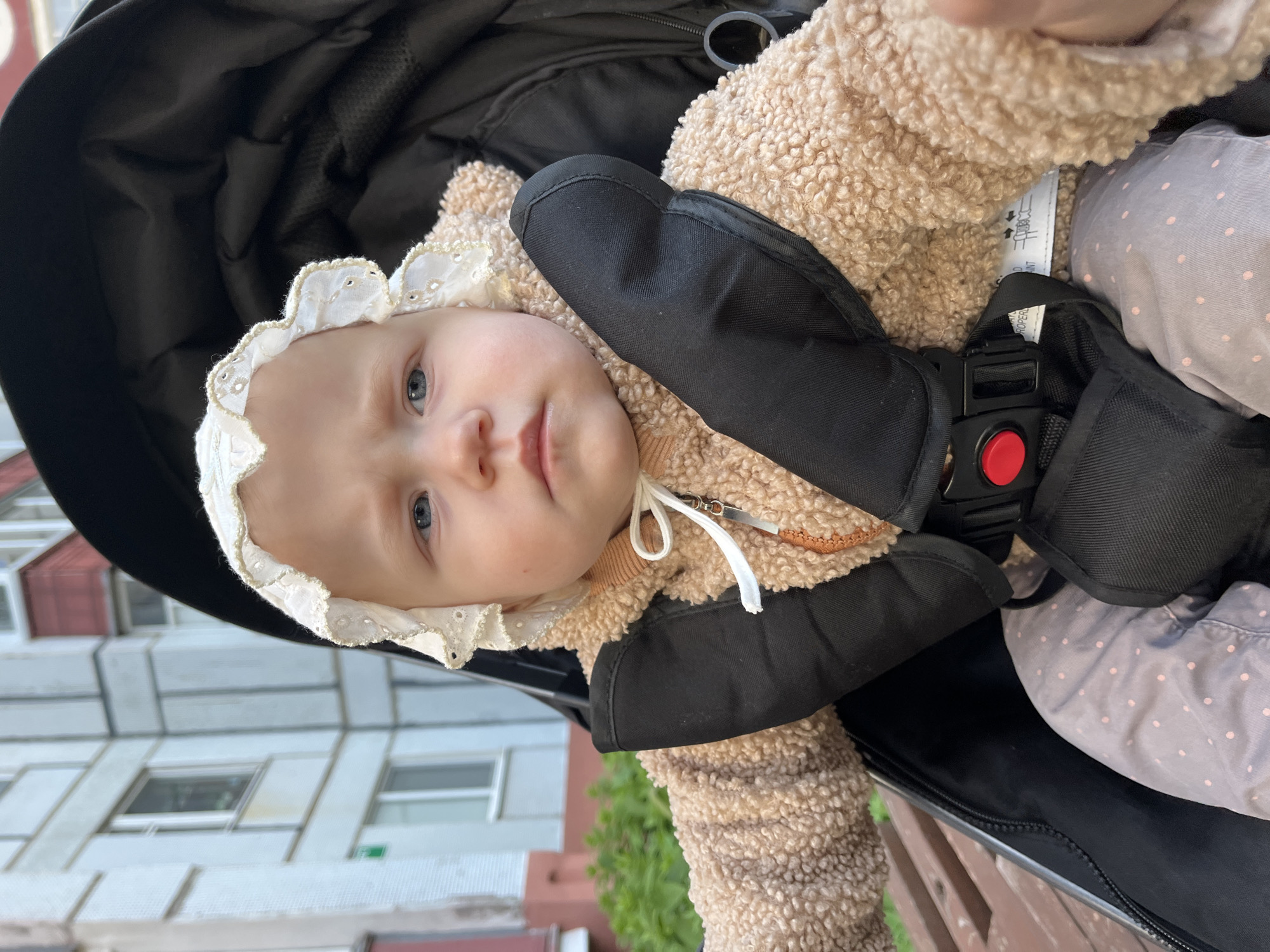 A baby is in a black stroller. The baby is wearing a white bonnet, a light brown curly fleece jacket, and black mittens attached to the jacket with cords. A safety harness secures the baby in the stroller. The building and surrounding greenery are viewed behind the baby. Based on the building construction, this is in the city of Chaikovsky, Russia.