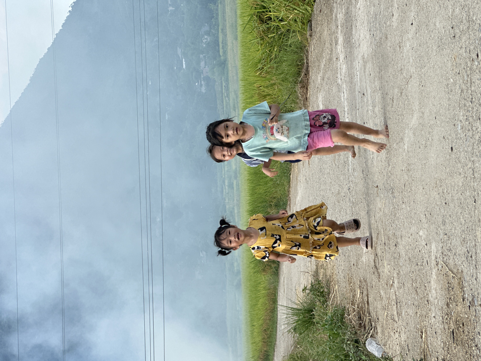 Three young girls are depicted running on an unpaved path in Gia Hội, Vietnam. The girl on the left, in the foreground, wears a yellow dress with a black and white cat pattern, has her hair in pigtails, and is wearing pink sandals, smiling with an open mouth. A second girl is partially visible behind and to the right of the first, wearing a striped shirt and also smiling while running. The third girl, in the foreground on the right, wears a light blue t-shirt with a colorful animal graphic and pink shorts featuring Minnie Mouse, and is running barefoot with a neutral expression. The path is dirt or gravel, flanked by sparse green vegetation, and a discarded clear plastic bottle lies on the left side of the path in the foreground. Beyond the path, extensive green fields, likely rice paddies, stretch horizontally. The background features large, hazy blue-grey mountains with scattered buildings visible on their lower slopes. Multiple dark power lines traverse the pale, overcast sky.