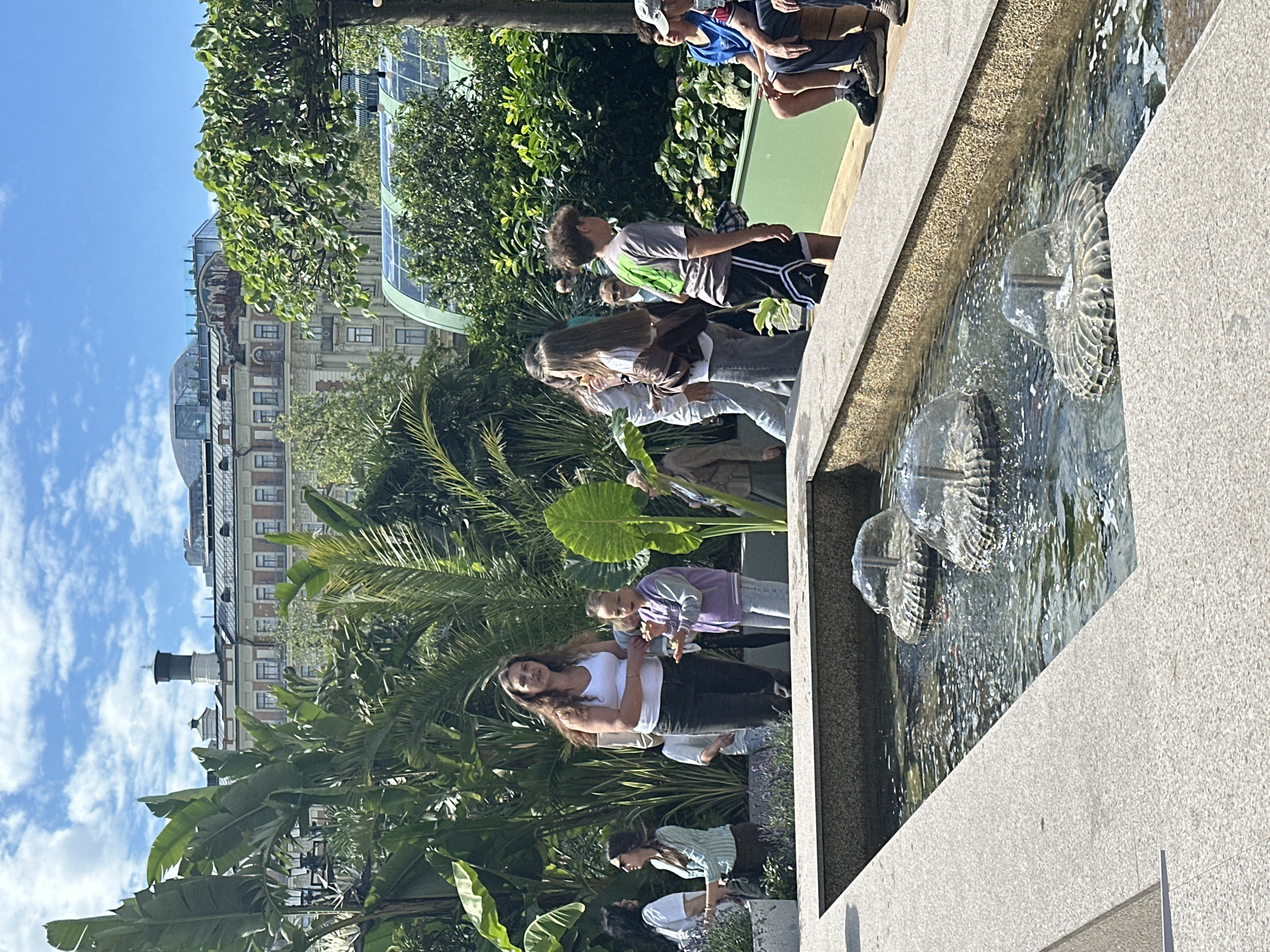 The image captures a sunny day scene in a lush, urban garden setting. In the foreground, a rectangular fountain with three water jets is visible. The water appears clear, with light rippling on its surface. The fountain is bordered by a light-colored stone ledge.
Behind the fountain, several people are scattered throughout the garden. On the left, a woman in a white tank top and dark jeans stands with a group of people. To her right, a young girl in a purple jacket holds something small in her hands. Further back, a diverse group of individuals are seen walking and standing amidst dense foliage. Tropical-looking plants with large leaves dominate the mid-ground, creating a verdant backdrop.
In the background, a series of buildings are visible, suggesting an urban environment. The architecture of the buildings appears classic, with multiple stories and windows. One building has a distinctive tower with a dark cap. The sky is blue with scattered white clouds.
The overall impression is of a lively, public garden space, likely a botanical garden or a well-maintained park within a city. Based on the architectural style of the buildings and the presence of lush, curated greenery, this scene is consistent with the Moscow Botanical Garden of the Moscow State University, or a similar high-profile garden within the city. The people appear to be enjoying a leisurely outing, with some observing the fountain and others interacting amongst themselves.