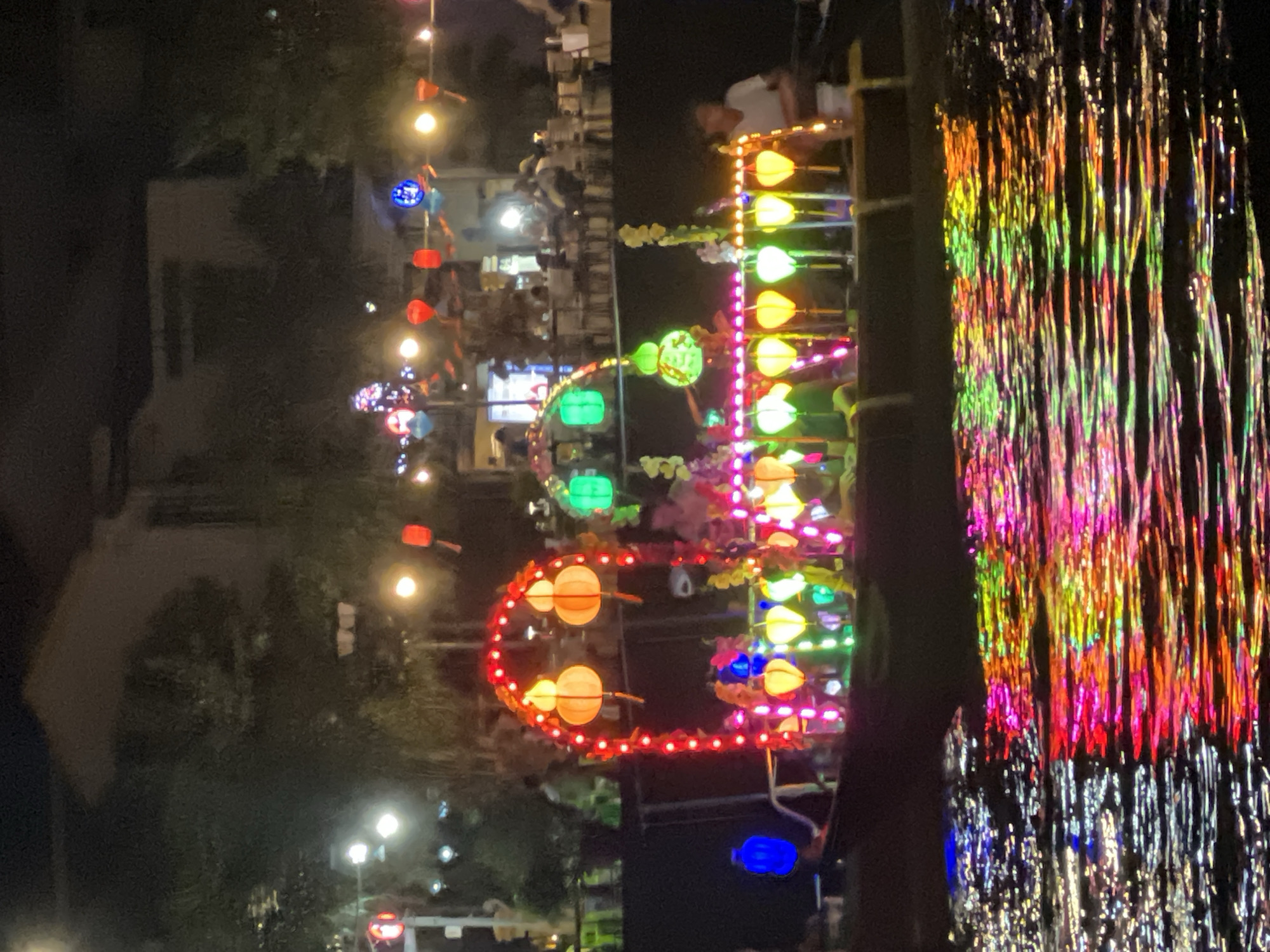 The image depicts a vibrant night scene along the Thu Bon River in Hoi An, Vietnam, during a lantern festival. A boat, heavily adorned with colorful lanterns and illuminated strings of lights, is the central subject. The boat's structure features an arched canopy decorated with red lights and two large, round orange lanterns hanging from it. Below this, a multitude of smaller lanterns in various shapes and colors – including yellow, green, and pink – are displayed along the boat's railing. The water in the foreground reflects the dazzling array of lights, creating a mesmerizing pattern of reds, oranges, greens, and blues. In the background, the dark silhouettes of trees and buildings are visible, with scattered lights suggesting other illuminated structures and possibly more boats. A few indistinct figures are present on the boat and on the riverbank. The overall atmosphere is festive and magical, characteristic of Hoi An's renowned lantern displays.