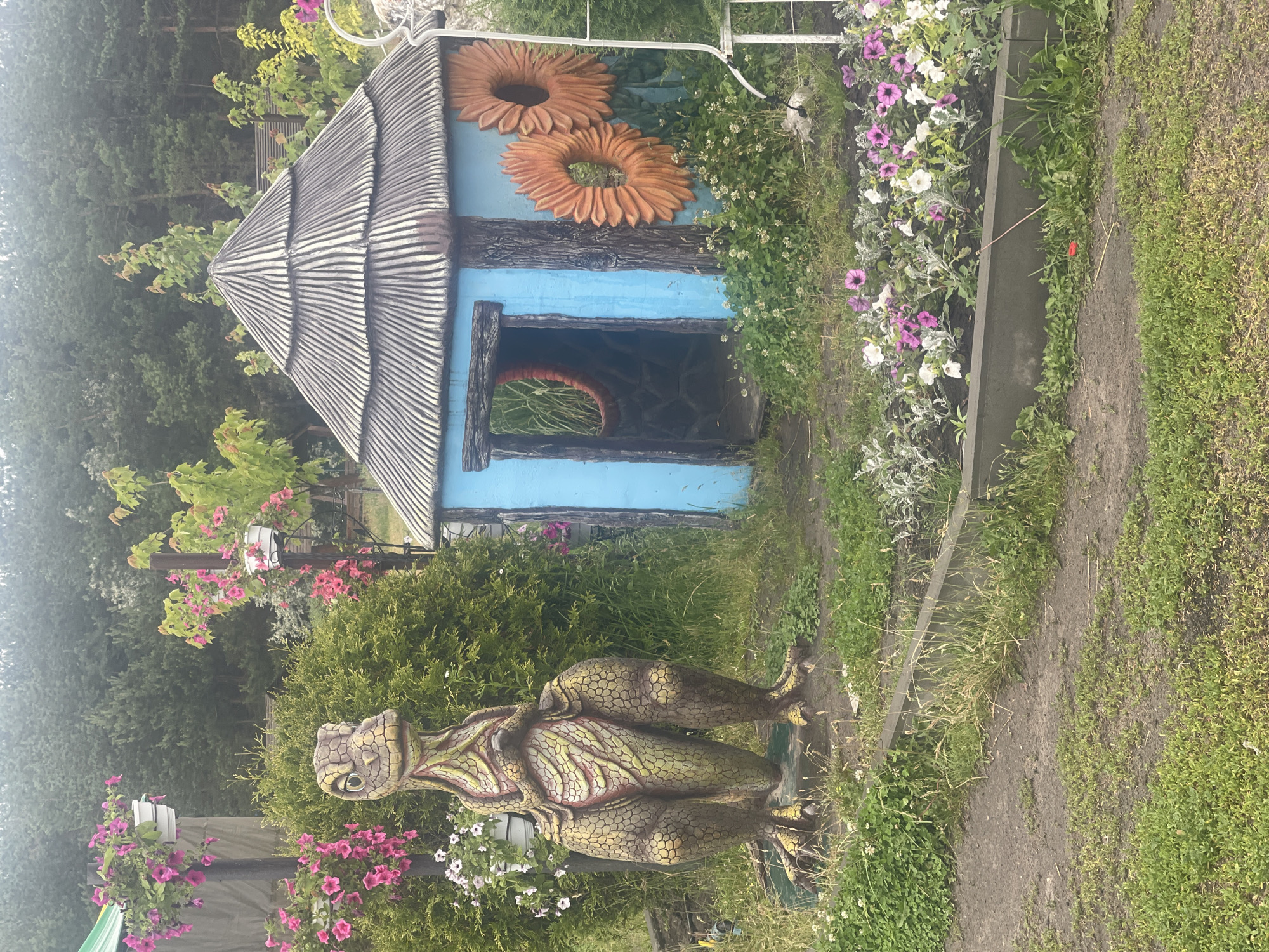 The image depicts a playful outdoor scene, likely in a garden or park setting. In the foreground, a statue of a dinosaur stands prominently, rendered in earthy tones with textured scales. To the right, a small, whimsical blue playhouse with a thatched roof is situated. The playhouse is adorned with large, orange sunflower decorations on its side. Surrounding these features are various plants and flowers, including pink petunias in hanging baskets and a flower bed with purple and white blooms. The background consists of dense green trees, suggesting a natural or semi-rural environment. The ground is covered with grass and dirt paths. The overall atmosphere is cheerful and inviting, evoking a sense of fantasy and childhood amusement. The image appears to be taken during the daytime under overcast skies.