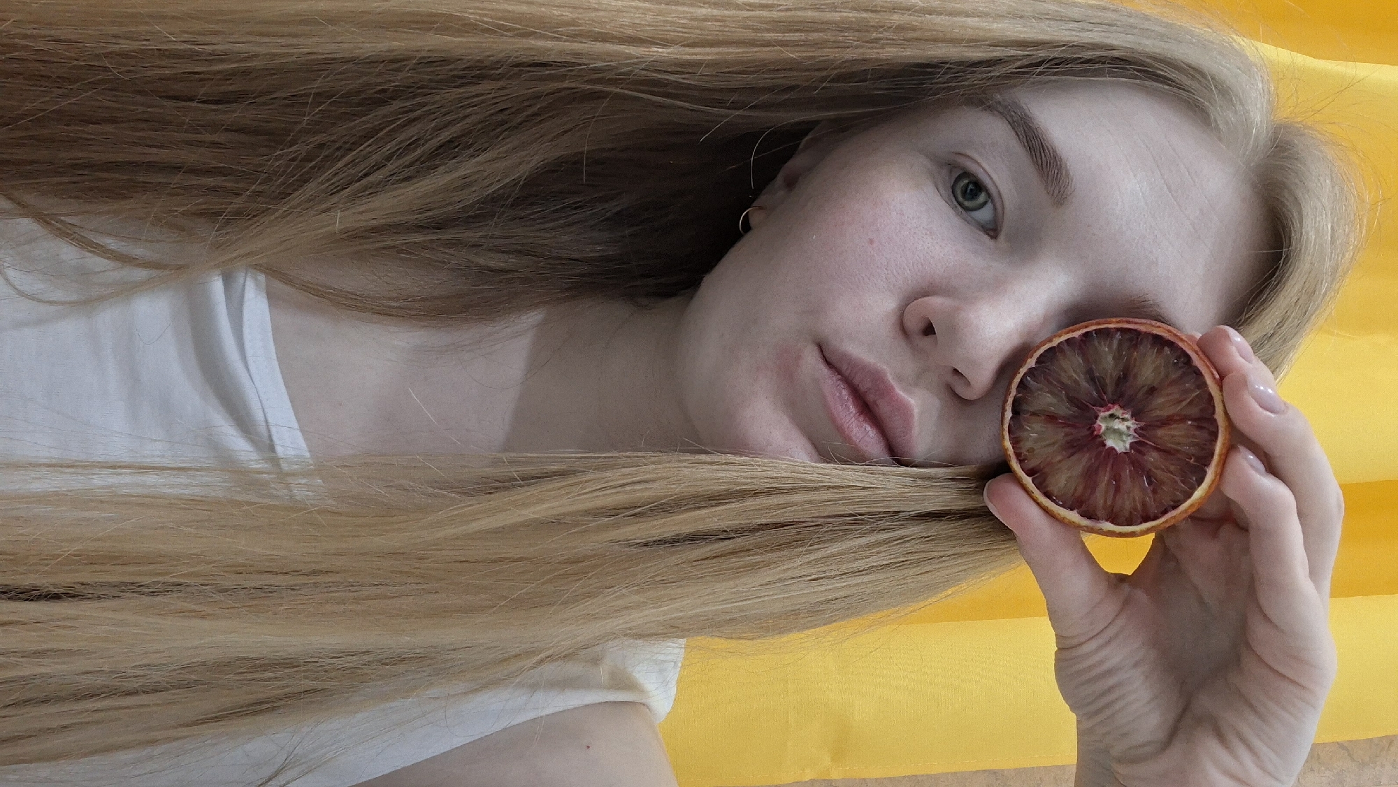 A woman with long, light blonde hair is lying down with her head turned to the side. She is holding a dried blood orange slice in her right hand near her face. She has light skin, green eyes, and is wearing a white top. The background is a solid yellow color. The location in the city of Chaikovsky, Russia is unknown.
