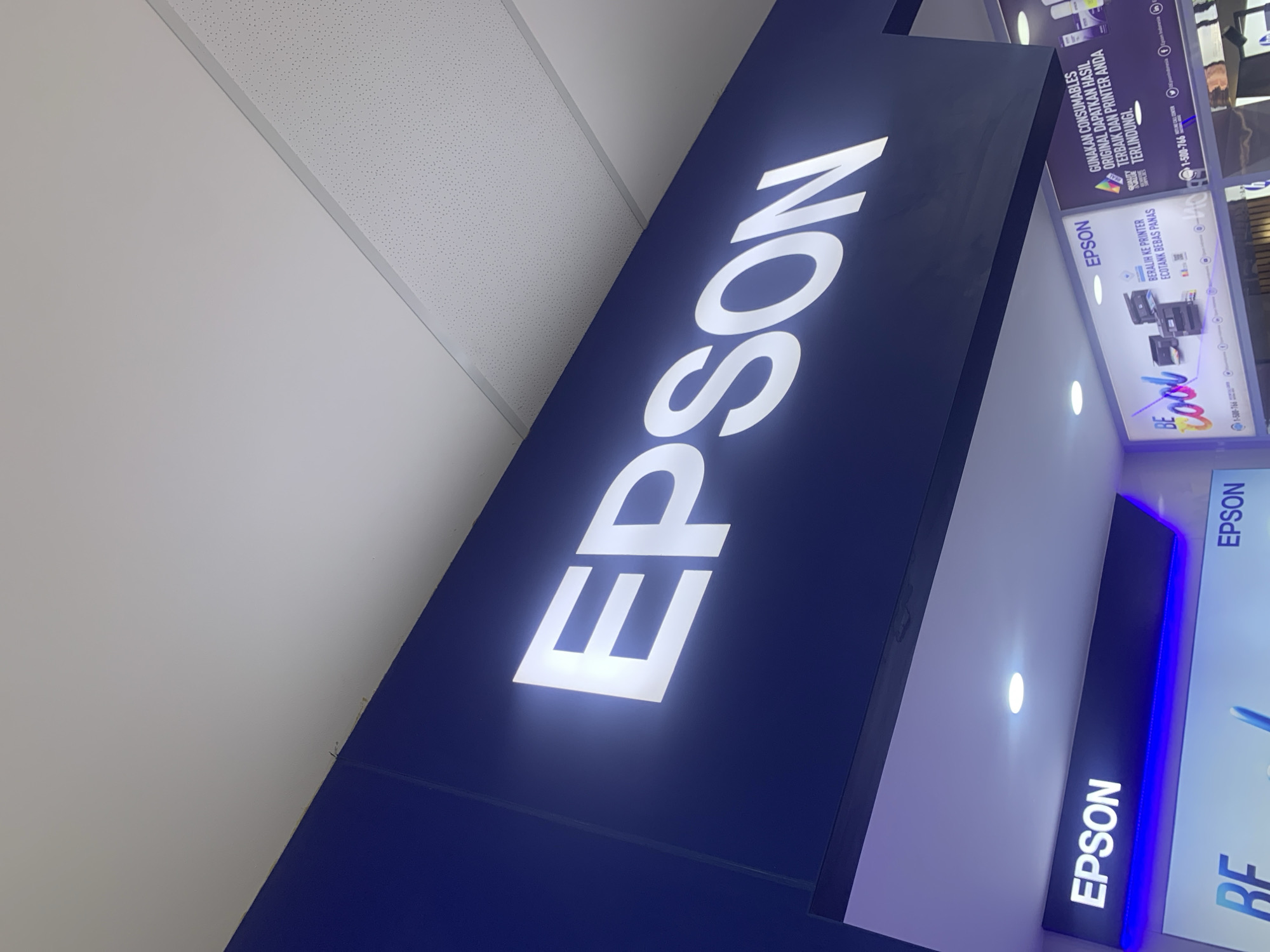 This image shows a sign for "EPSON" in large, illuminated white letters against a dark blue background. The sign is positioned above a display of Epson products. Below the main Epson sign, another illuminated sign also displays the "EPSON" logo in white letters, with blue LED lighting underneath. Further down, there are other displays featuring Epson printers and text in what appears to be Malay or Indonesian, promoting Epson's products and services. The scene is indoors, likely in a retail store or showroom, with a white, speckled ceiling visible above the signs. The image appears to have been taken at a slight upward angle, looking towards the signs. While the city of Palembang, Indonesia, is mentioned in the prompt, there are no specific visual cues within the image that confirm this location.