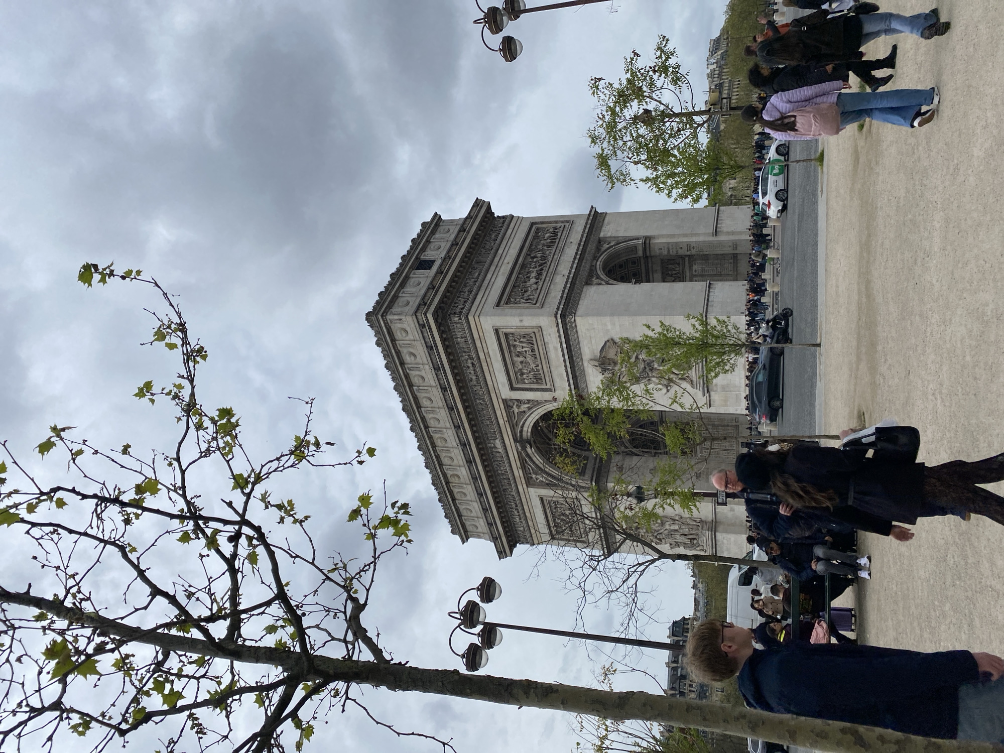 The image captures the Arc de Triomphe in Paris, France, on an overcast day. The monument's architectural details are visible, including its arches and sculpted reliefs. People are seen walking and standing around the base of the Arc de Triomphe. Cars and other vehicles are parked along the road next to the Arc. Trees with sparse spring foliage frame the monument. A lamppost with globe lights is in the foreground, adding to the urban setting.