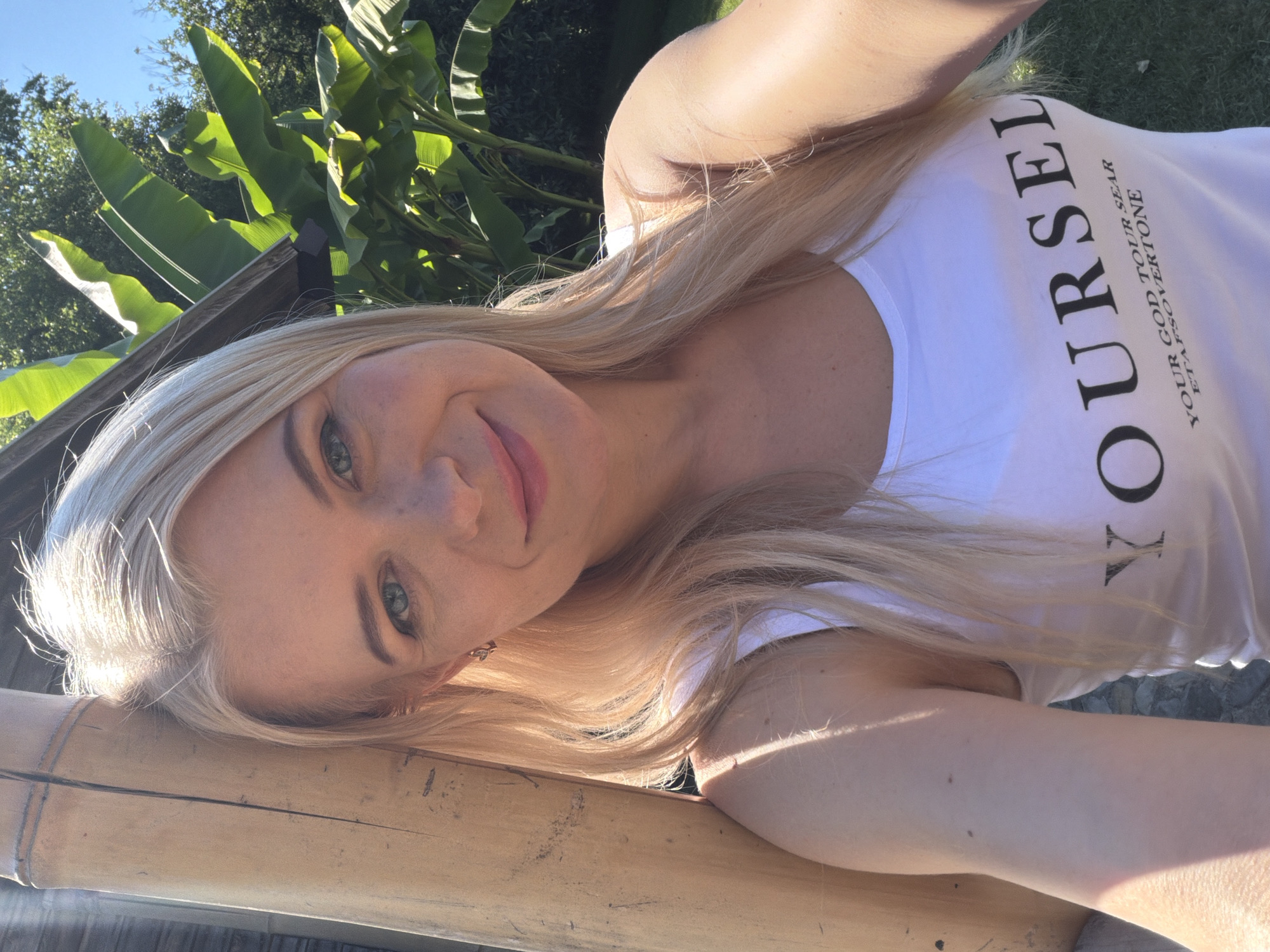 This media file features a blonde woman outdoors, with lush green foliage in the background. She is leaning on a wooden surface, possibly a railing or bench, made of bamboo. The sunlight casts strong shadows, suggesting it is daytime, likely morning or afternoon. The woman is looking towards the camera, with a slight smile. She is wearing a white t-shirt with black text printed on it. The text reads "YOURSELF" in large, bold letters, with smaller text below that says "YOUR GOD TOUR SEAR ETAPSOVERTONE." The overall mood appears relaxed and cheerful. The greenery suggests a warm climate, consistent with a location like Sochi, Russia, especially during warmer months.