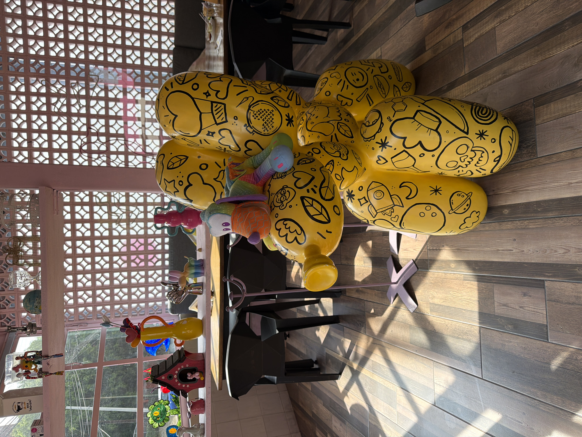 This media file captures an interior scene, likely a vibrant cafe or shop, characterized by eclectic and whimsical decor. The main subject is a large, bright yellow sculpture positioned towards the right side of the frame. This sculpture is composed of several balloon-like, elongated forms clustered together, and is extensively decorated with black line-art doodles including faces, hearts, stars, leaves, a rocket, a planet, a skull, food items like a taco and banana, and various abstract patterns. A colorful, segmented, worm-like plush toy with blue, green, orange, and purple sections is draped over one of the yellow forms. The sculpture rests on a purple cross-shaped base.
The flooring consists of wood-look tiles laid in a plank pattern, exhibiting variations in shade. Strong sunlight streams into the space, casting distinct shadows on the floor from the sculpture and nearby furniture, indicating it is daytime and sunny outside. In the background, there are black chairs and tables.
To the left, a light-colored, possibly pinkish-white, decorative lattice screen forms part of the wall or a partition. Beyond this lattice, windows reveal glimpses of greenery outside. A shelf or ledge running along the left edge of the image displays a variety of decorative objects, including small colorful figurines, a miniature globe, glassware, a red birdhouse, a yellow vase with colorful embellishments, and other ornamental trinkets.
The overall atmosphere is cheerful and artistic. While many doodles are visible on the yellow sculpture, no distinct, readable text is apparent, beyond occasional letters like "OH" as part of a drawing. The scene is static, without any visible people or ongoing activity, focusing solely on the decorative elements of the interior space. The location context of Puebla, Mexico, aligns with the colorful and artistic aesthetic.