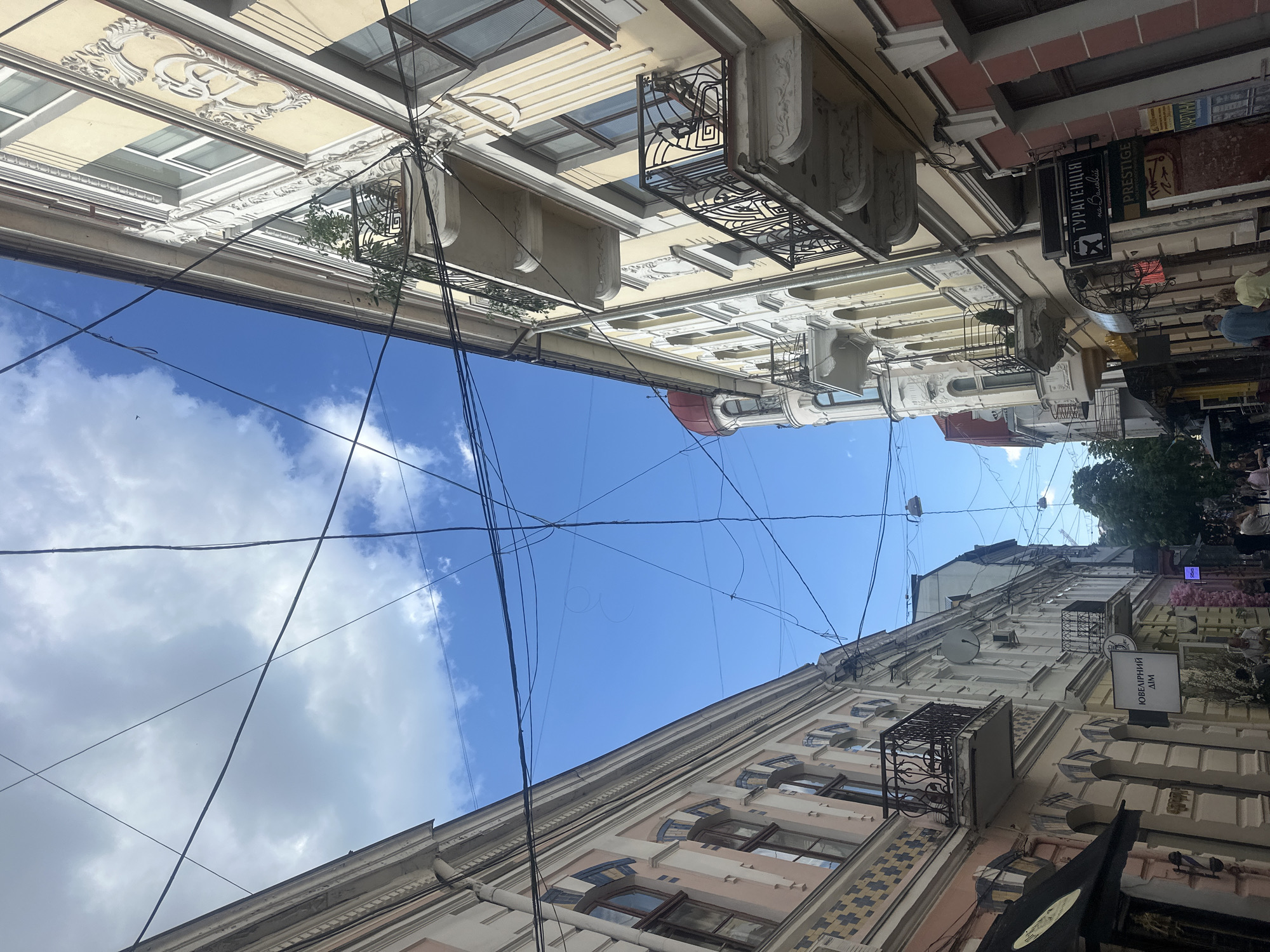 This media file shows an upward-facing view of historic buildings lining a narrow street, likely in Ternopil, Ukraine. The architecture features ornate facades with decorative molding, balconies with wrought-iron railings, and numerous windows. Overhead, a network of tangled electrical wires crisscrosses a bright blue sky dotted with fluffy white clouds. The scene suggests a sunny day, possibly in the late morning or afternoon, given the clear sky and ambient light.
The buildings appear to be a mix of residential and commercial spaces. Several signs are visible on the facades, though the text is mostly illegible or in Cyrillic script. One sign reads "ПРЕСТИЖ" which translates to "Prestige". Another sign appears to say "ТУРАГЕНЦІЯ" which means "Travel Agency". There are also hints of people on the street below, partially visible at the bottom right of the frame, suggesting it is a public thoroughfare. The overall impression is one of a vibrant, old-world European city street.