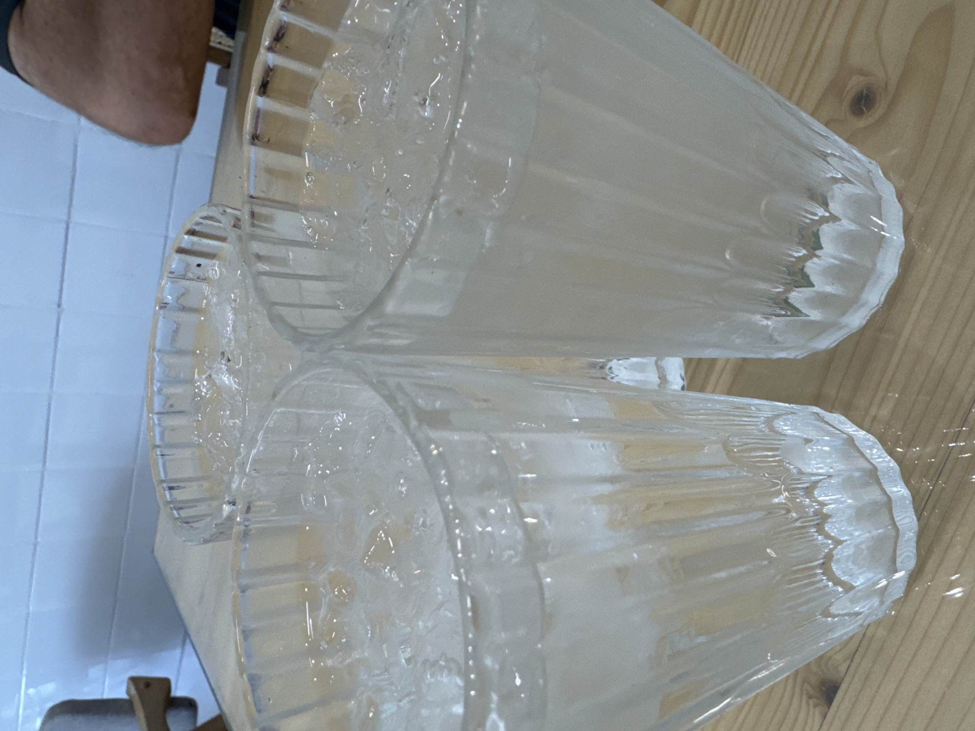 The image shows three glasses filled with ice and a light-colored liquid, likely water or a beverage, placed on a wooden table. The glasses have a ribbed texture. In the background, there is a tiled wall and a person's arm and shoulder are visible on the right side. The scene is indoors, possibly in a restaurant or bar. There is no identifiable information to determine the location in Kuala Lumpur, Malaysia.