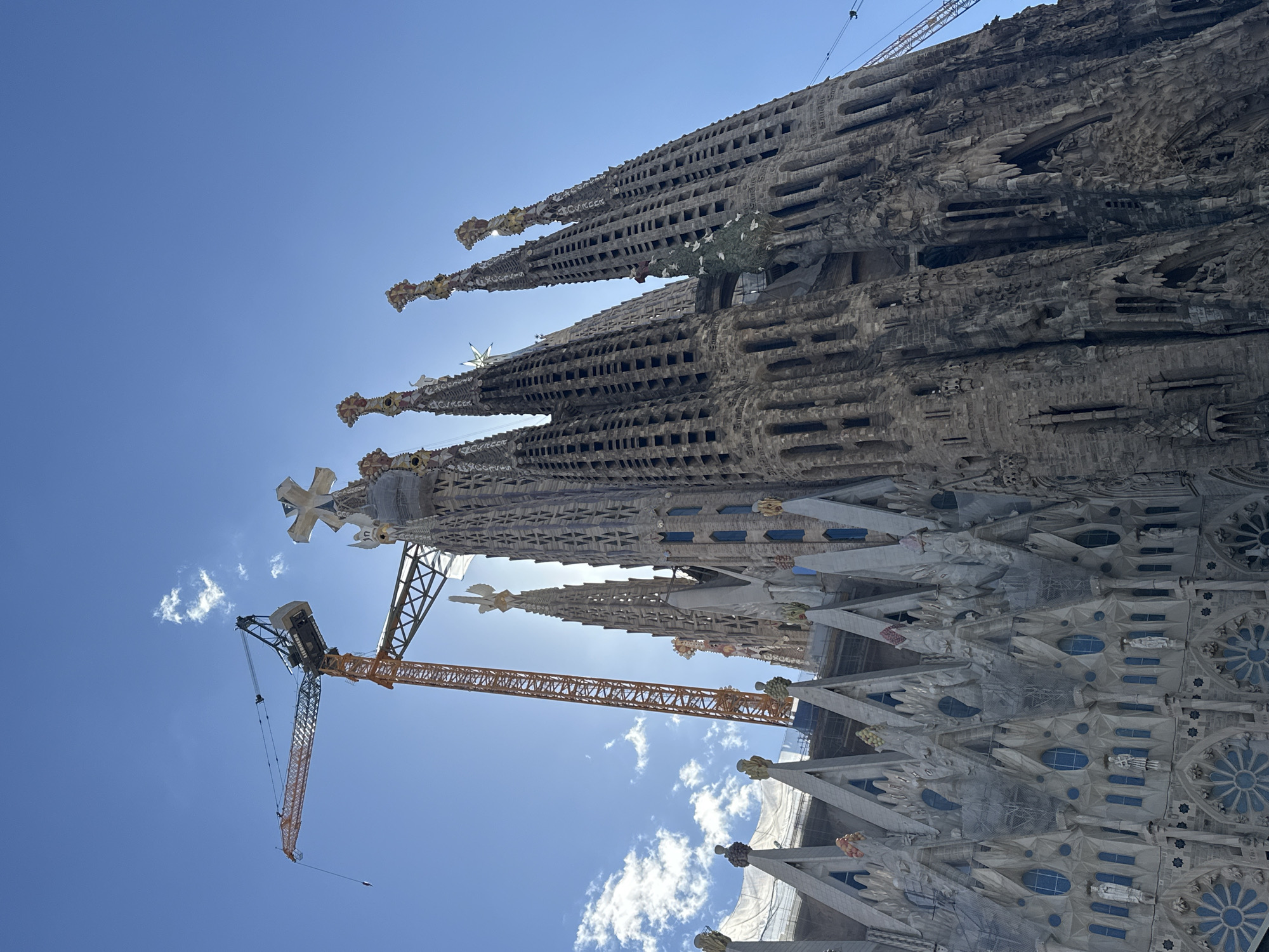 The image depicts the Sagrada Familia basilica in Barcelona, Spain, viewed from a low angle. The spires of the basilica dominate the frame, showcasing intricate stonework and details. A construction crane is positioned near the basilica, indicating ongoing work on the building. The sky is bright blue with scattered clouds. The architecture is complex, with numerous openings and carvings visible. The scene is well-lit, with the sun behind the basilica creating a slightly backlit effect.