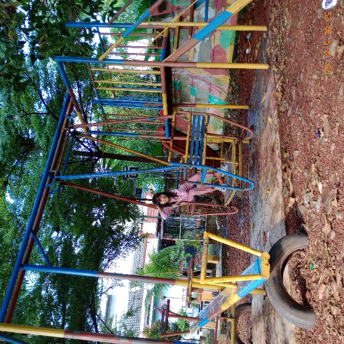 The image shows a colorful playground located in Panongan, Indonesia (latitude: -6.275, longitude: 106.551). It features a swing set, a slide, and a seesaw. A young girl is sitting on the swing, smiling at the camera. She is wearing a pink shirt and blue pants. The swing set is made of metal and has a blue and yellow color scheme. The slide is yellow and blue, and the seesaw is made of wood. The playground is surrounded by trees and grass. The ground is covered in dirt and leaves. The image is taken from a low angle, looking up at the playground. The sky is blue and there are clouds in the background.