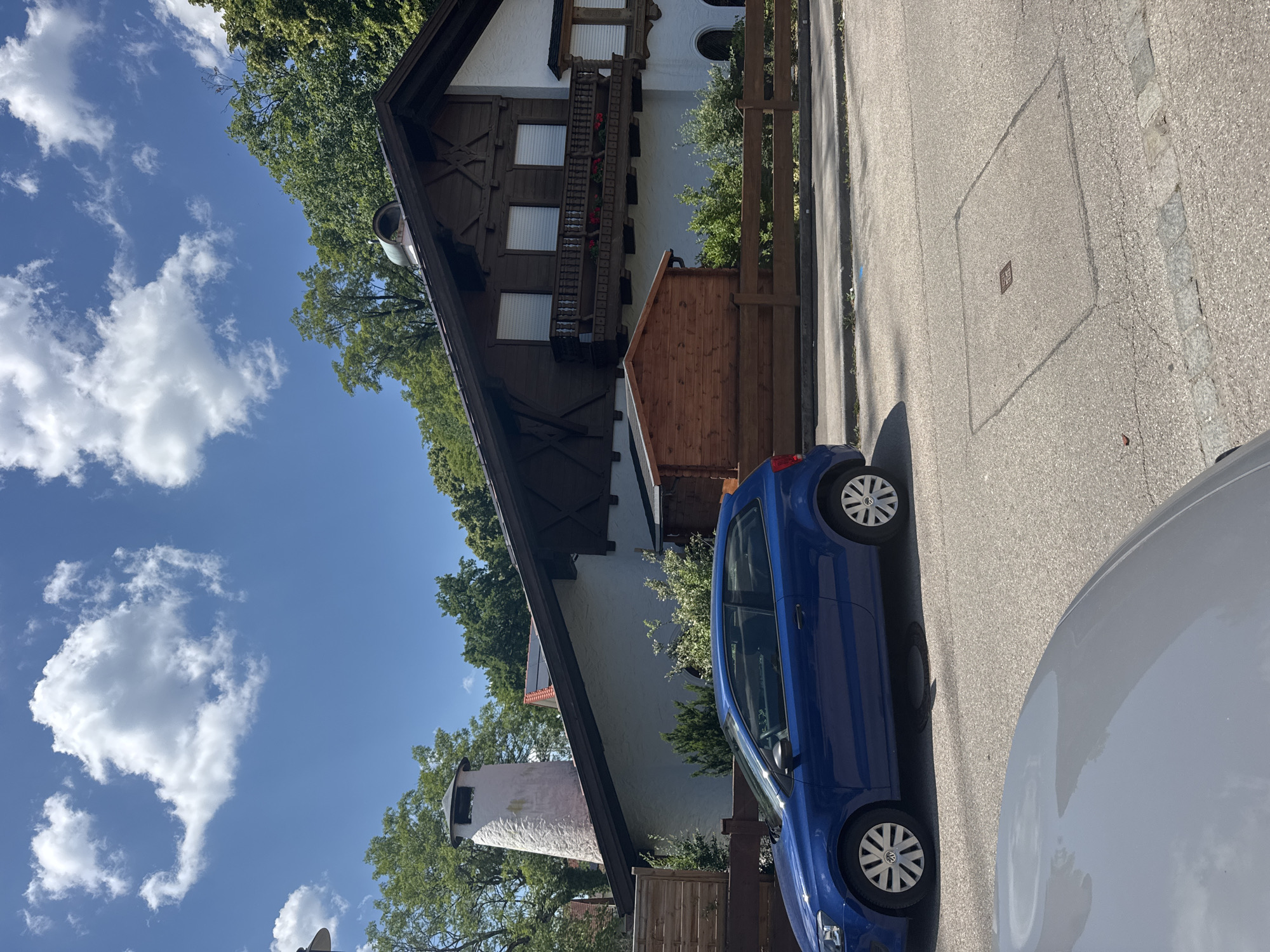 On a clear, sunny day in Höhenkirchen-Siegertsbrunn, Germany, a blue Volkswagen Polo hatchback is parked on the right side of a residential street, facing left. The car is situated on asphalt, with a grey car partially visible in the foreground, also parked on the street. A wooden fence runs along the property line behind the blue car.
The property features a large, multi-story house with traditional Bavarian architecture, characterized by dark wooden trim and white stucco walls. A prominent, conical white chimney stands to the left of the house. A small, wooden shed is visible in front of the house, just behind the fence. Trees line the property and the hillside behind the house, indicating a semi-rural or suburban setting. The sky is blue with scattered white clouds. There are no people visible in the image.