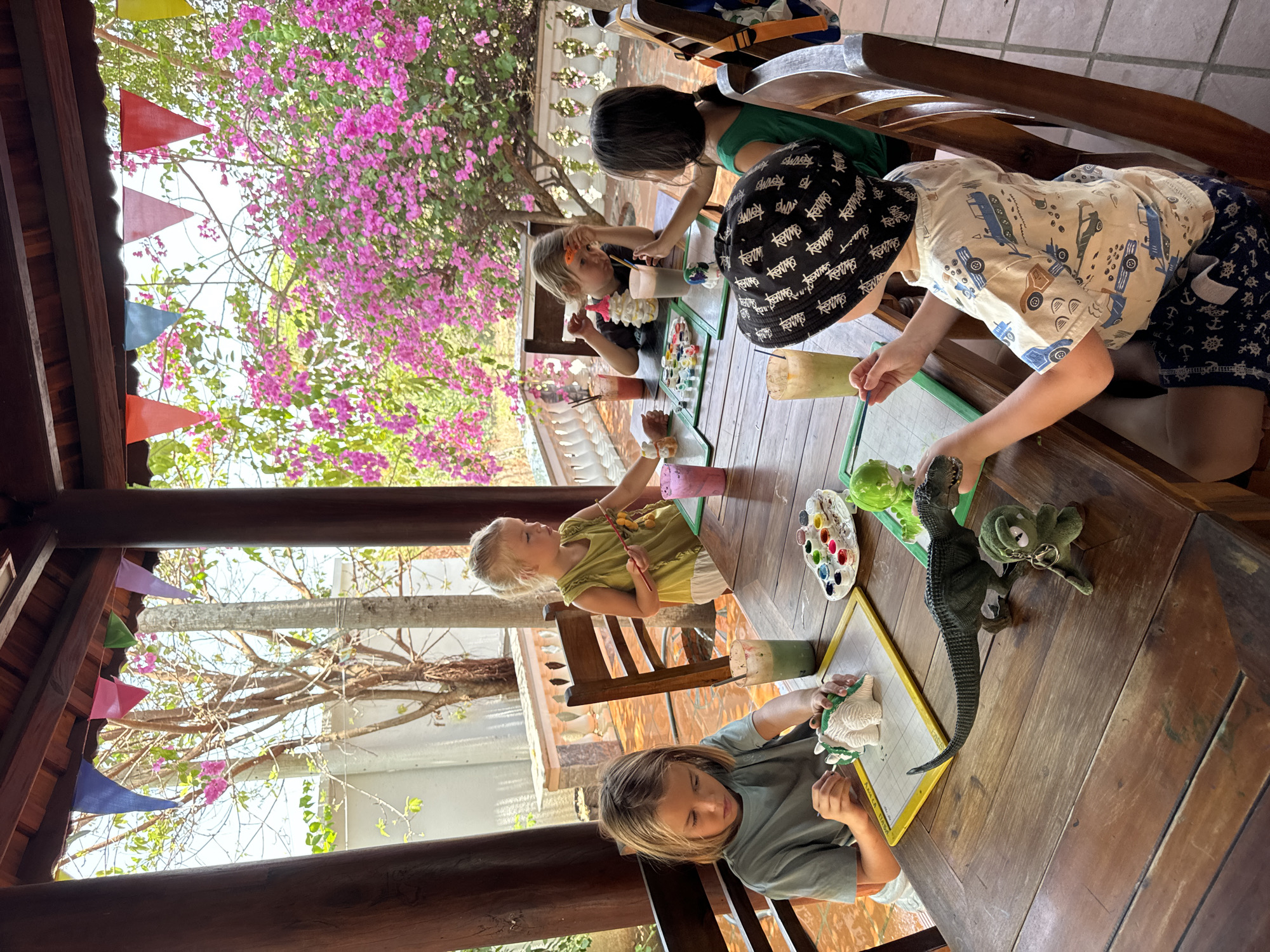 The image shows five children sitting around a wooden table outdoors in Mũi Né, Vietnam. They are engaged in an art activity, painting ceramic dinosaur figurines. Each child has a small palette of paints and paintbrushes. Some of them are actively painting, while others are watching or assisting. The background features a vibrant display of pink bougainvillea flowers and colorful pennant banners. The setting appears to be a veranda or covered patio area.