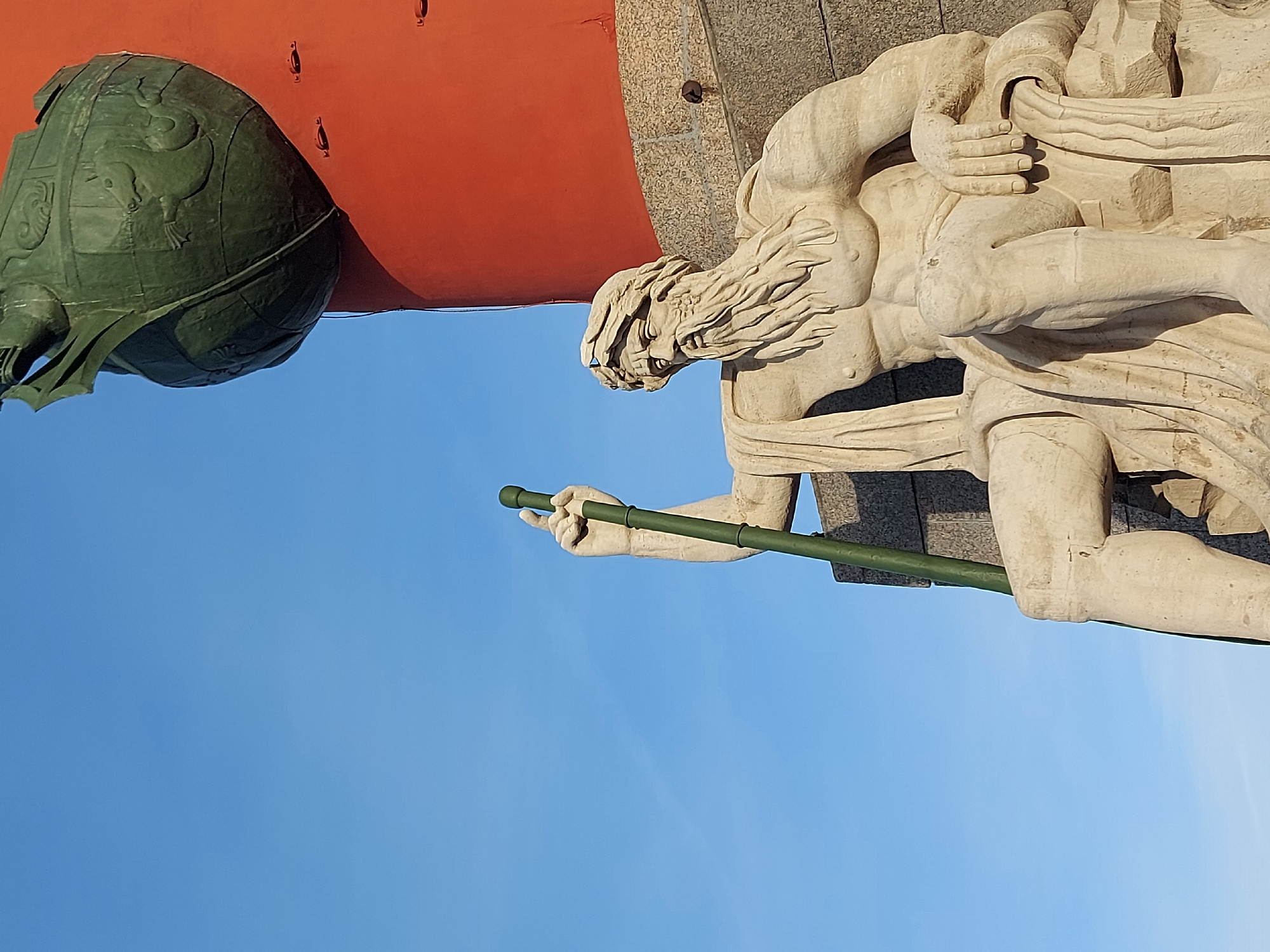 The image shows a low-angle view of a portion of the Alexander Column in Saint Petersburg, Russia. The column is partially visible, featuring a red-colored section and a green sphere-shaped element adorned with animal reliefs. Below these are the figures of a monument. In the lower right quadrant of the image, there is a statue of a man holding what appears to be a long, green spear or staff. The background is a clear, blue sky.