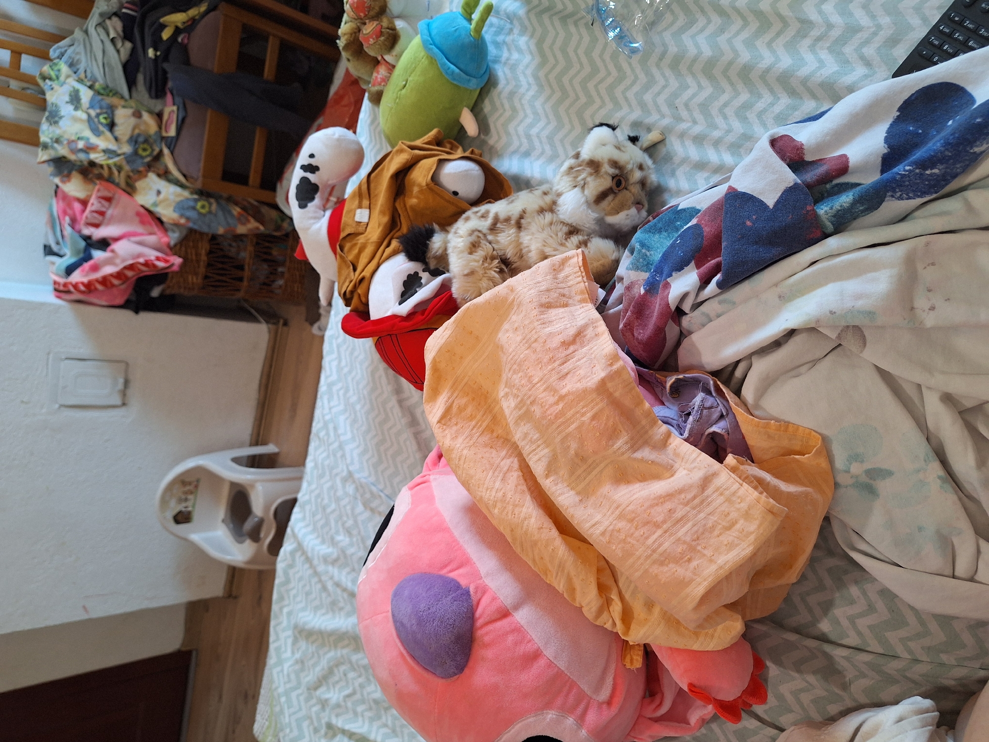 In a room in Blagoevgrad, Bulgaria, a bed is covered with a collection of stuffed animals and various fabrics. A pink stuffed character with a purple ear is prominent, partially obscured by an orange textile. To its right, a plush toy dog with white and brown markings lies on a mustard-colored garment. Further to the right, a green cylindrical stuffed toy with blue accents is visible. In the background, a white child's potty chair is situated near a wall. A woven basket and a wooden chair piled high with colorful fabrics and clothing complete the scene. The overall impression is one of a disarranged personal space.