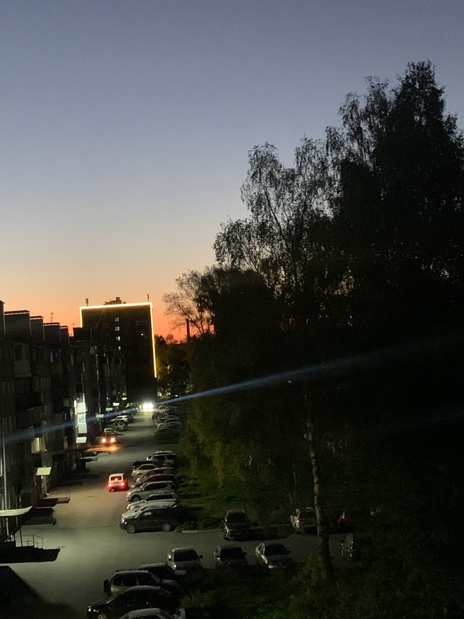 An outdoor, high-angle, dusk shot shows a residential area with buildings, parked cars, and trees. The sky transitions from a pale blue at the top to a warm orange and pink near the horizon, indicating sunset or sunrise. A tall, dark building in the center is outlined with bright yellow lights. To the left, apartment buildings with visible windows are lined up. A street or parking lot with numerous cars, some with headlights on, runs through the scene. Dense trees, mostly silhouetted against the sky, occupy the right side of the frame. The overall impression is a quiet evening in an urban environment.