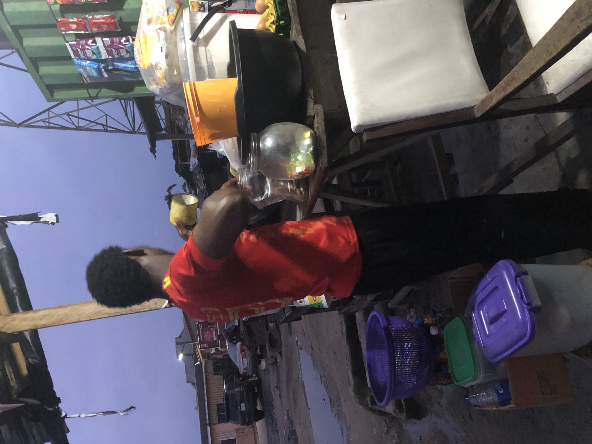 The image depicts an outdoor scene at dusk, likely in Ado, Nigeria, where a person wearing a red t-shirt and black pants is preparing something on a wooden table. The table holds several items, including a glass jar filled with colorful candies, an orange bucket, a large black basin, and a clear plastic container. In the background, a green shipping container is visible, along with a metal tower structure. Further in the distance, buildings and vehicles suggest an urban or semi-urban environment. To the left of the table, a purple basket and various containers of drinks are placed on the ground. A white chair with a wooden frame is positioned in the foreground to the right. The overall impression is of a street-side vendor preparing goods for sale.