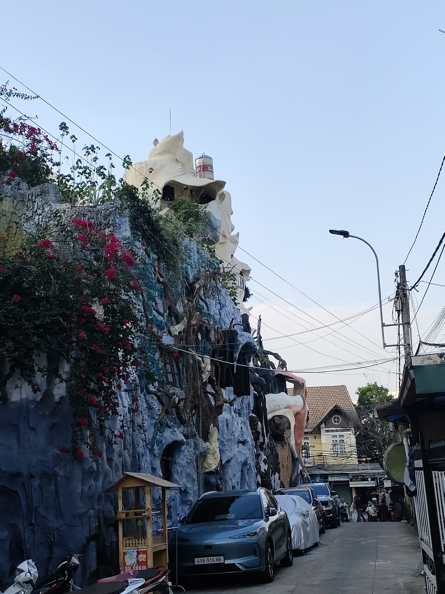 The image depicts a street in Da Lat, Vietnam, featuring the "Crazy House" (Hang Nga guesthouse) prominent on the left. The unique architecture of the building mimics natural forms, with its sculpted walls, tree-like structures, and organic shapes. The building is adorned with various plants and flowers. In the foreground, several parked cars are visible along the street. A small wooden structure stands near the building, adjacent to parked motorbikes. Further down the street, a yellow building with a round window is visible. In the background, a group of people is gathered. Power lines and a street lamp are visible overhead. The sky is a clear light blue.