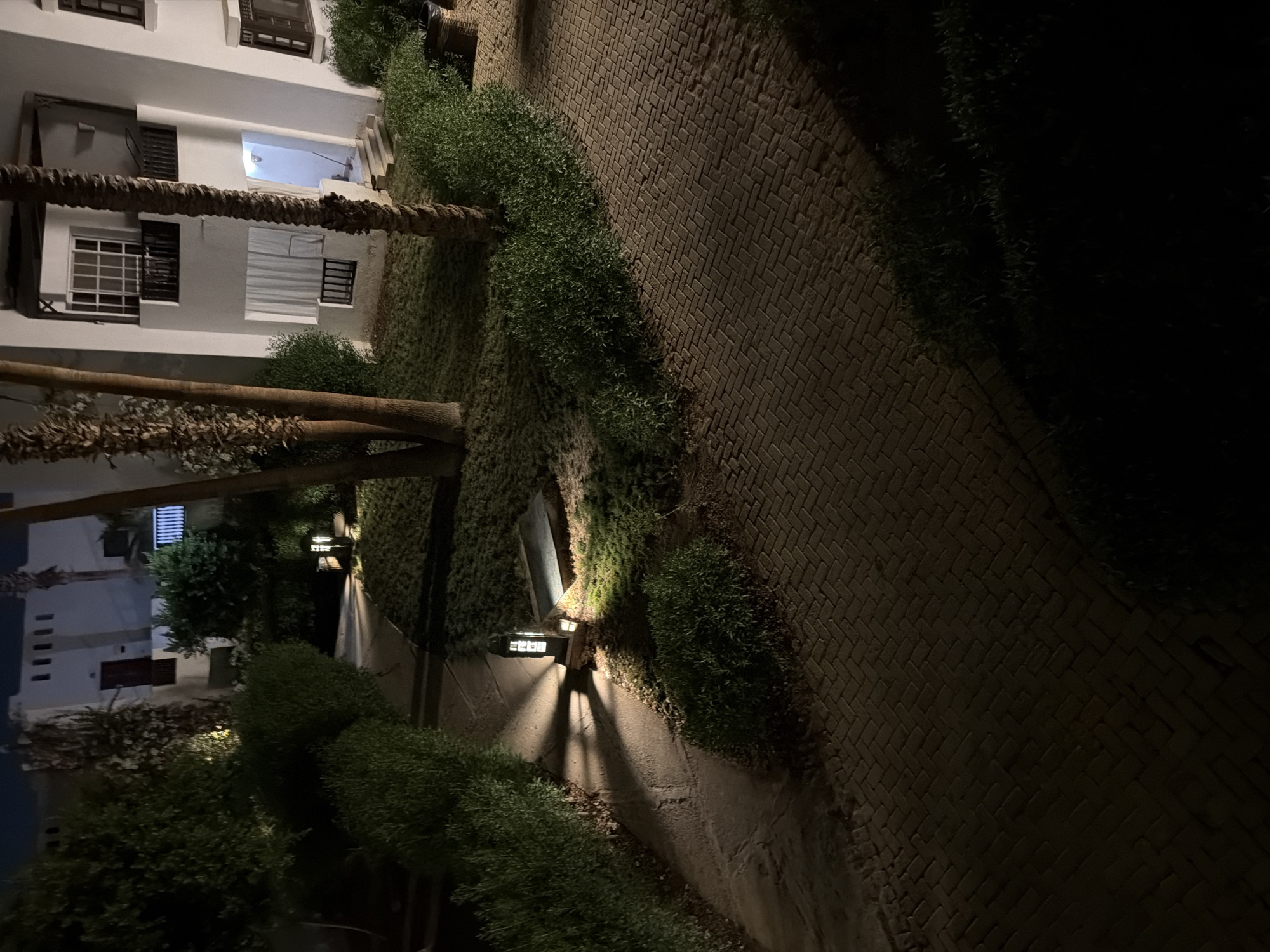 The image depicts a nighttime scene in Sharm El Sheikh, Egypt, featuring a paved walkway and landscaped grounds illuminated by a lantern. A white, multi-story building with balconies and windows is visible in the background, partially obscured by palm trees. The walkway is constructed of interlocking bricks, and the surrounding area is covered in grass and low-lying bushes. The lantern emits a warm light, casting shadows across the path and onto the grass. The overall impression is one of a well-maintained outdoor space in a resort or residential area.
