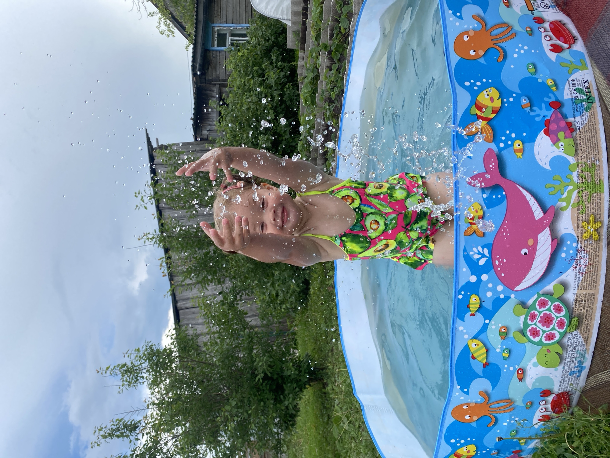 A young girl is splashing in a small, inflatable kiddie pool filled with blue water. The pool has a colorful underwater scene printed on its sides, featuring octopuses, fish, a whale, and a turtle. The girl, wearing a swimsuit with an avocado print, is smiling and has her arms raised, with water droplets flying around her. The pool is situated on a grassy area in a backyard. In the background, there are trees, a wooden fence, and a rustic wooden house with a blue-framed window. The sky above is cloudy. The location is identified as Pesnevo, Russia.