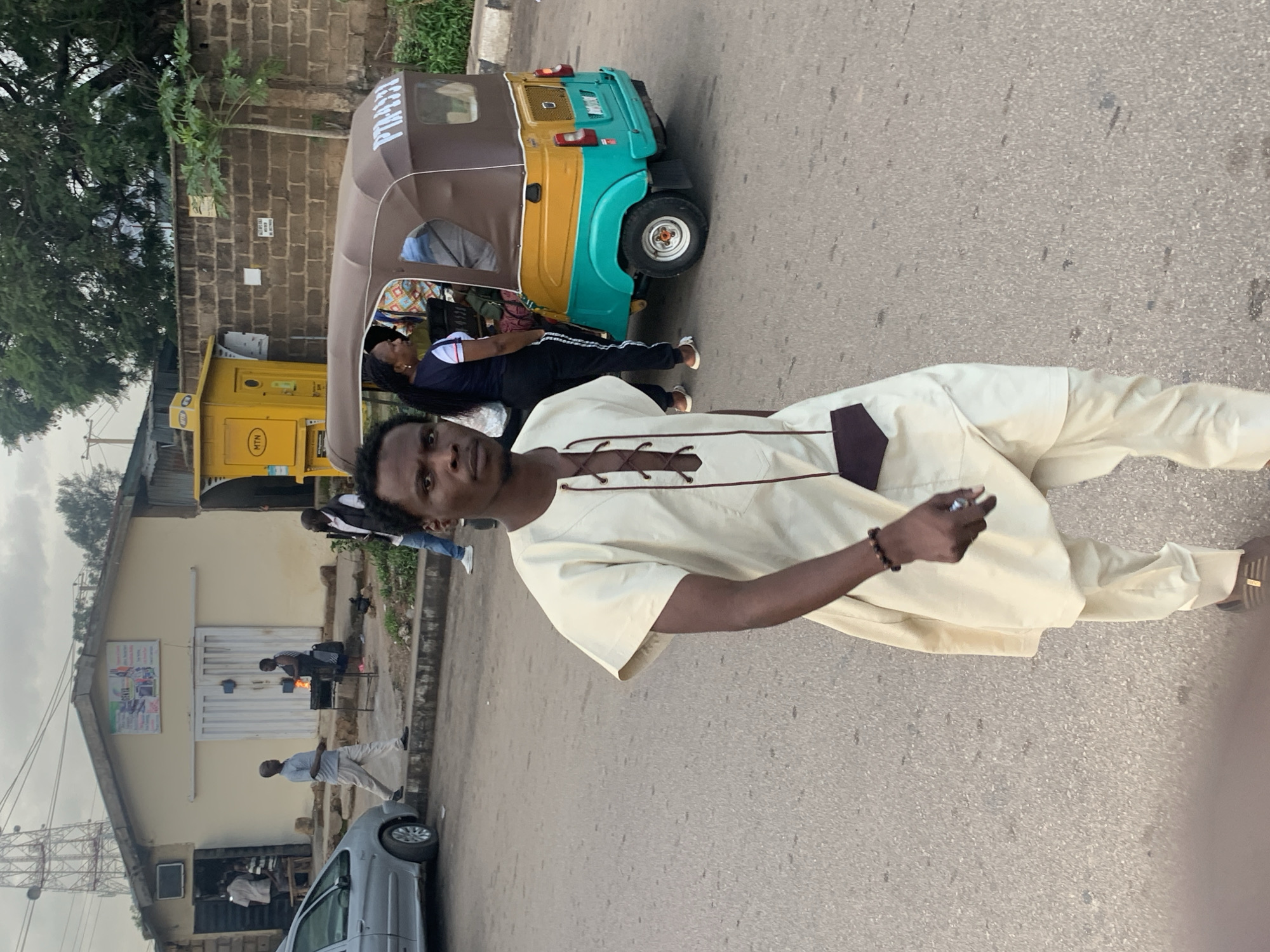 In Barakin Lerawi, Nigeria, a young Black man wearing a light-colored traditional outfit walks on a paved street. He is the primary subject of the image, positioned centrally and facing forward, with a slight blur indicating movement. He has a dark pouch-like object hanging from his neck and is wearing a beaded bracelet on his left wrist. In the background, a colorful auto-rickshaw, primarily yellow and teal, is parked on the side of the road. A woman wearing a dark top and white-striped pants stands near the auto-rickshaw. Further back, a yellow MTN booth is visible. To the left, a silver car is parked, and a building with a sign featuring multiple images is behind it. Several other individuals are present in the background, some walking and others standing near the building. The scene appears to be a street in a moderately populated area.