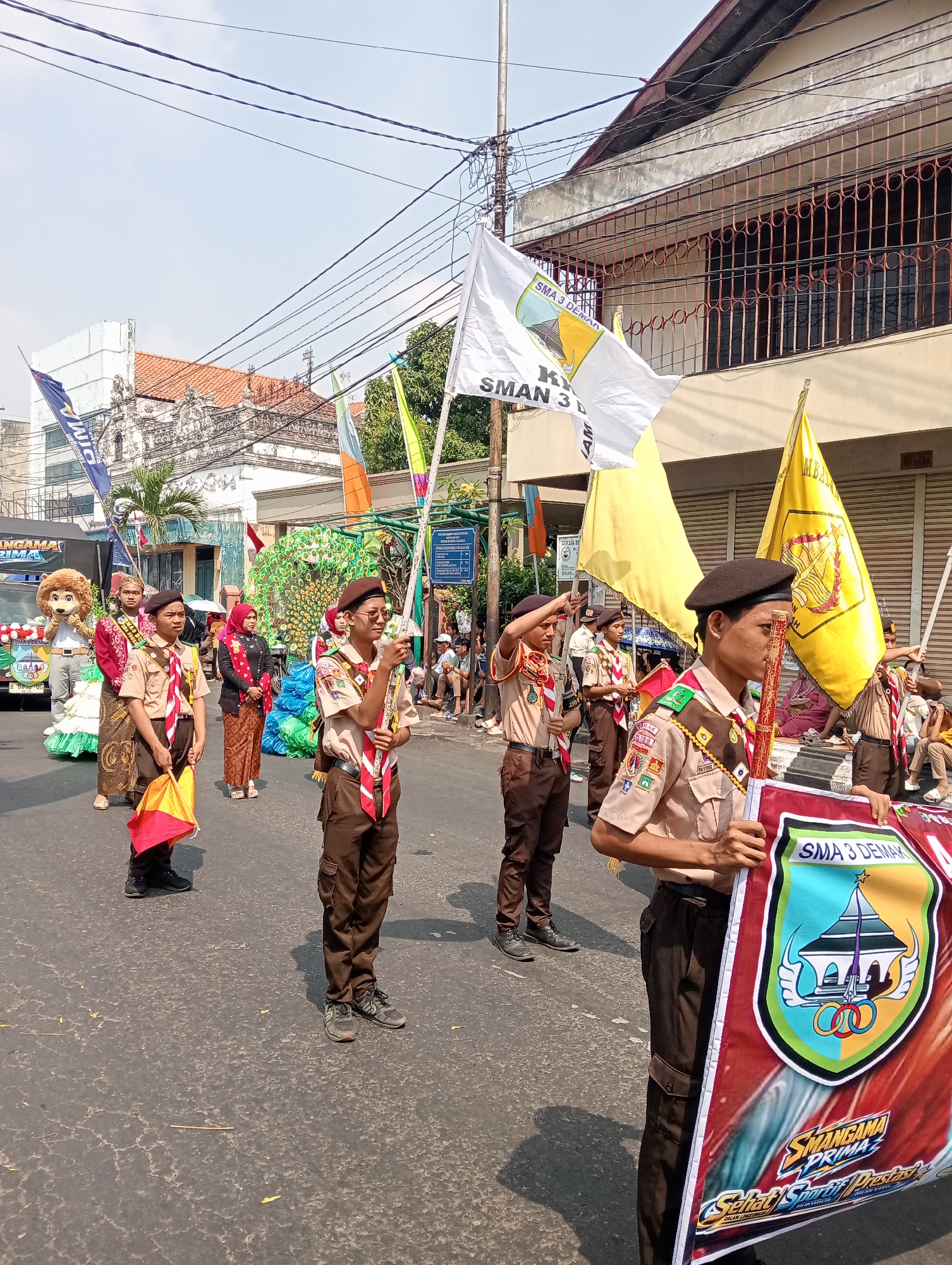 The image shows a parade in Bintoro, Indonesia. Several students in scout uniforms are marching down a street, holding flags and a banner. The banner in the foreground features a shield with a building and Olympic rings, along with text that appears to be in Indonesian. In the background, there are buildings, a peacock-themed float, and spectators watching the parade. The weather appears to be sunny.