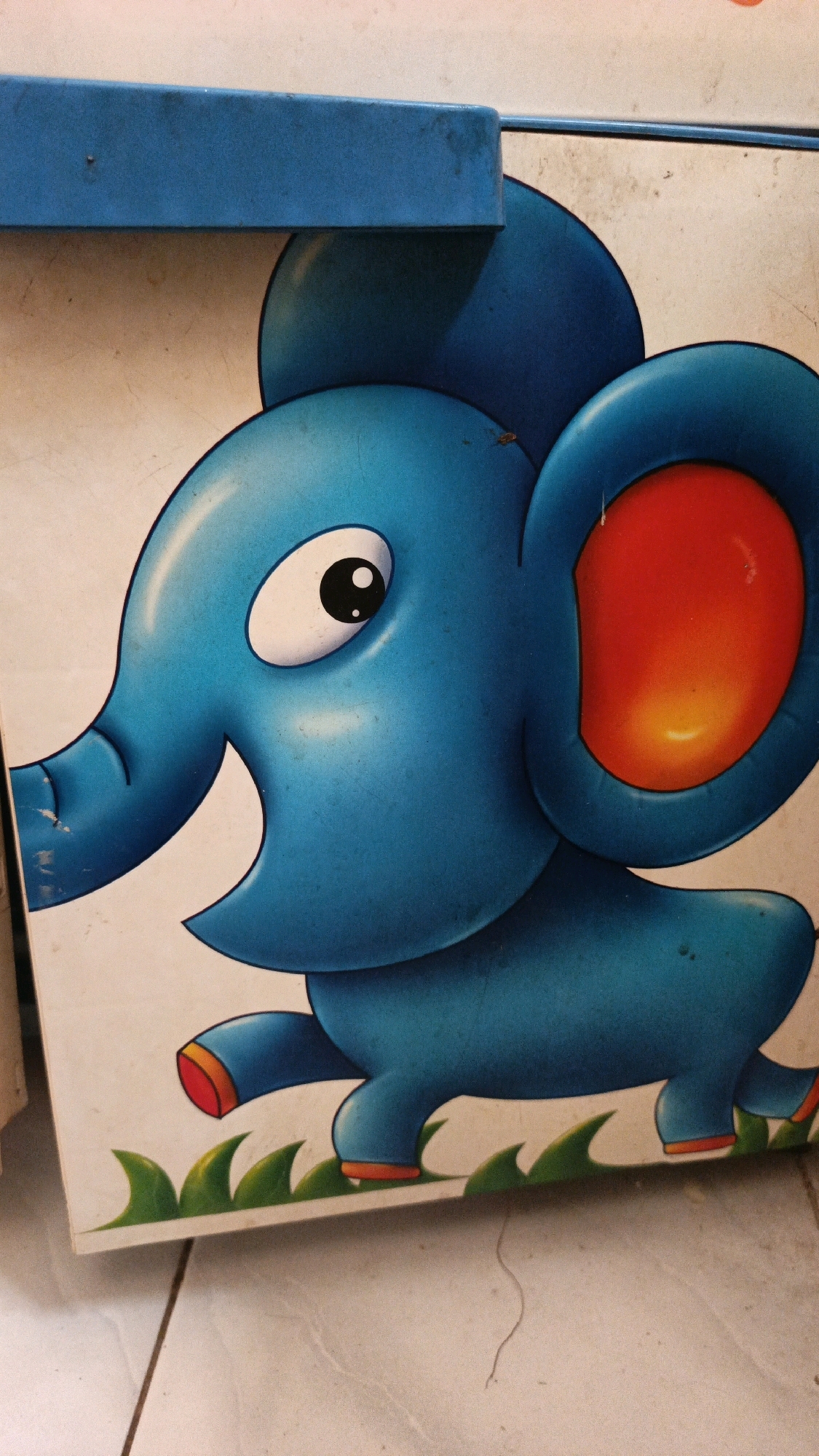 A cartoon illustration of a blue elephant is depicted on a white background. The elephant is shown in profile, facing left, with its trunk curled upwards. It has large, round ears with a bright red and yellow gradient inside, and a single, large eye with a black pupil. The elephant's feet are orange. Green, stylized blades of grass are painted at the bottom of the illustration. A blue, rectangular object is positioned along the top edge of the image, partially obscuring the elephant's head and ears. The scene appears to be a close-up shot of a graphic on a flat surface, possibly a wall or a piece of furniture. The lighting is even, and there are no discernible shadows that would indicate a specific time of day or natural light source. The environment is not clearly identifiable as being in North Kuta, Indonesia, as no specific landmarks or cultural elements are visible in the image.