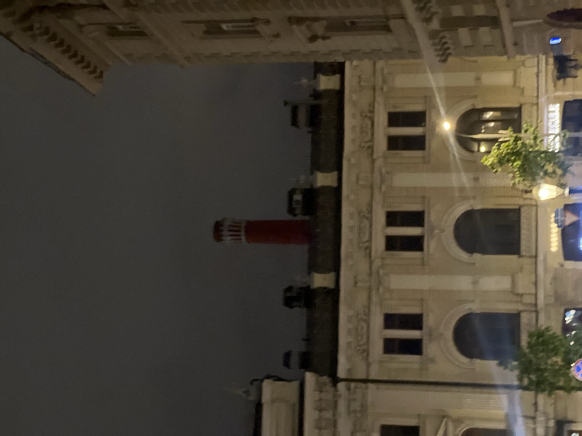Here is a description of the image:
The image shows a nighttime view of buildings in what appears to be Moscow, Russia. A large beige building with ornate architectural details is prominently displayed on the right side of the image. A red-and-white striped tower is visible in the distance towards the left of the building. The sky is a dark gray, suggesting it is night or a very overcast day. The scene is urban, and architectural details suggest a historical part of the city. There are trees planted in front of the building. Light illuminates parts of the building, indicating the presence of streetlights or internal building lights.