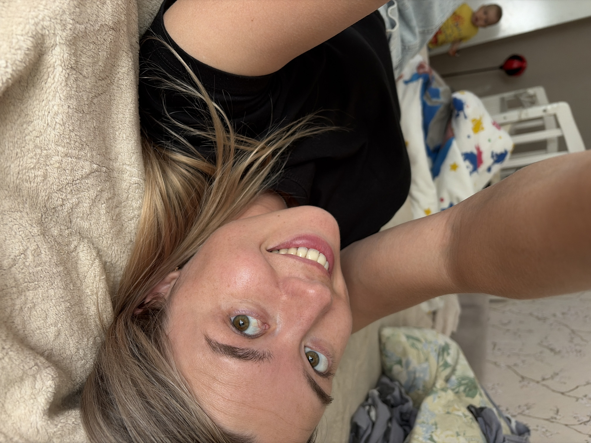 An overhead shot shows a woman lying on a beige, textured blanket, possibly a bed. She is wearing a black t-shirt and has blonde hair. Her head is tilted, and her eyes are looking upwards and to the left. The background is slightly out of focus, showing a child in a yellow shirt standing in front of a white crib. There are also some pillows and blankets visible in the background, with one featuring a pattern of horses and stars. The overall impression is a candid shot taken indoors, likely in a bedroom or nursery. The location is not discernible from the image.
