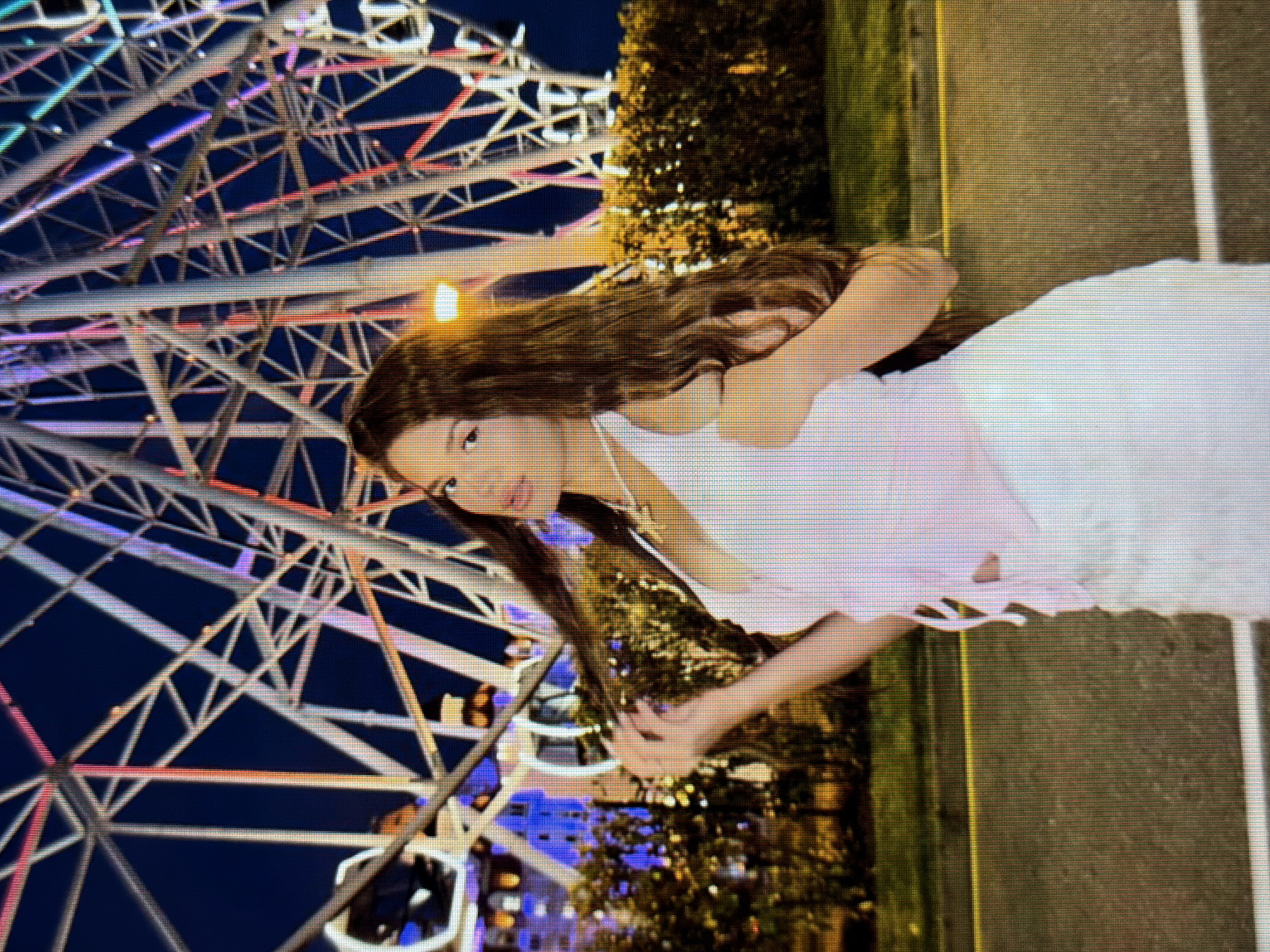 A medium shot shows a woman with long, dark, wavy hair posing in front of a Ferris wheel at night in Sirius, Russia. She is wearing a light pink halter top and a white skirt. Her left arm is bent at the elbow, and she is touching her hair with her left hand. The Ferris wheel, which is illuminated with colorful lights, is visible in the background. There are also trees and bushes behind the woman, and a road with a yellow line is in the foreground. The sky is dark blue, indicating it is nighttime.