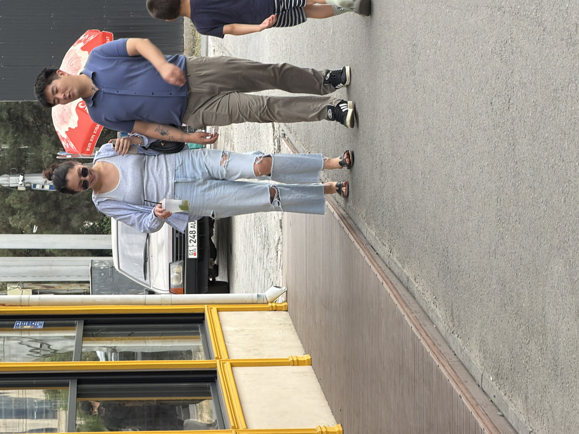 A man and a woman walk side-by-side on a paved surface. The woman, on the left, wears ripped light blue jeans, a grey tank top, and an unbuttoned light blue shirt. She has sunglasses on and holds a drink in her right hand. The man, on the right, wears khaki pants and a blue polo shirt. They are walking away from the camera and slightly towards the right. Behind them, a child in a dark shirt and striped shorts walks in the same direction. To the left of the couple, a building with a yellow trim and large windows is visible. A car, partially visible, is parked in front of the building. In the background, a red umbrella with white lettering is open. The scene appears to be outdoors in an urban setting.