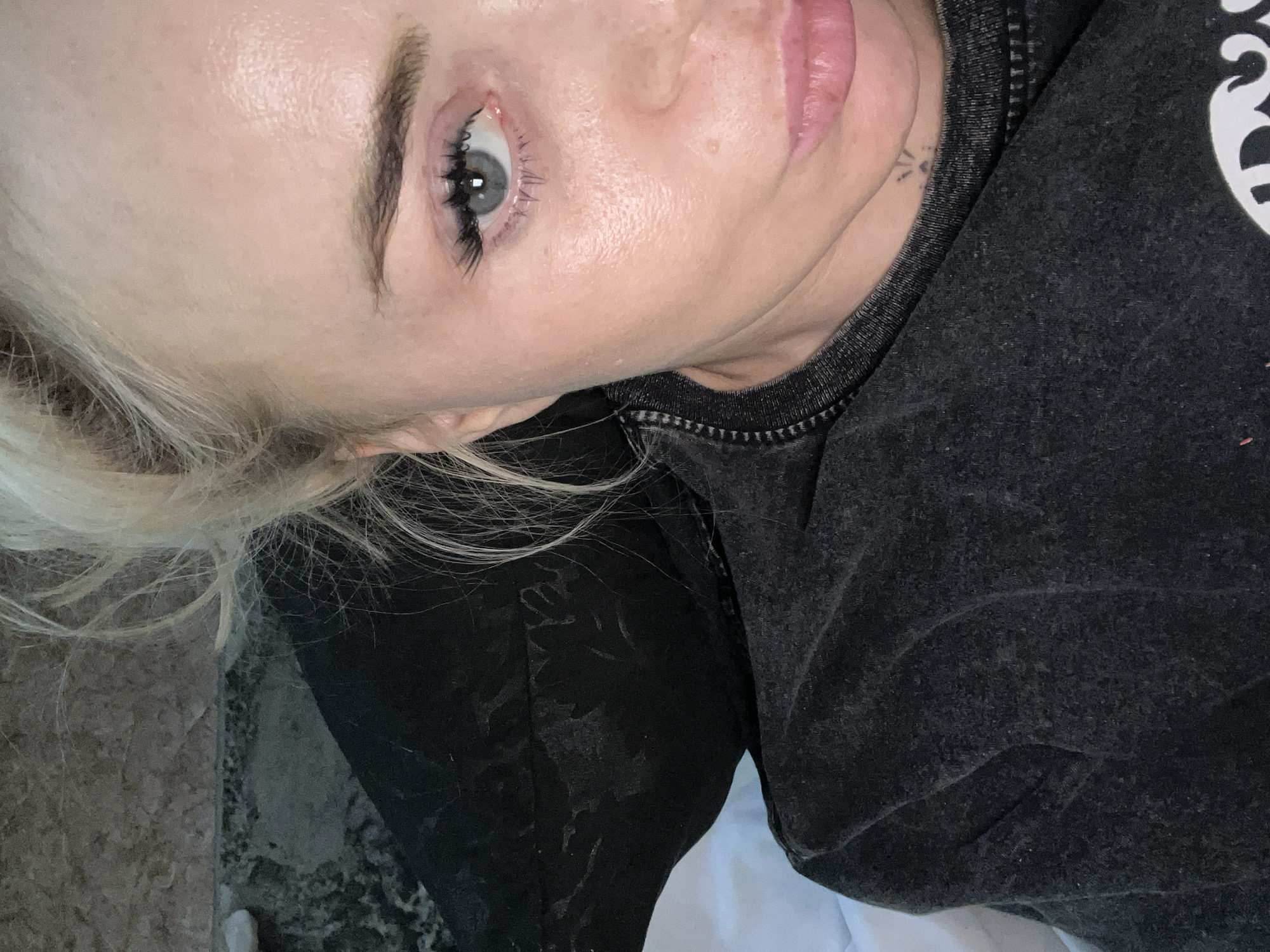 The image is a close-up, selfie-style photograph of a blonde woman's face and upper torso. She is wearing a dark gray t-shirt with a textured pattern visible on the fabric. Her hair is pulled back, with loose strands framing her face. The woman's eye, which is a light blue, is prominent, and she is wearing false eyelashes. Her skin appears to have a dewy or oily sheen. A small, subtle tattoo of a sunburst is visible on her neck, just below her jawline. The background is out of focus, but appears to include dark fabric and a textured surface that could be a wall or furniture. The scene is indoors, with no specific identifying landmarks of a city or location.