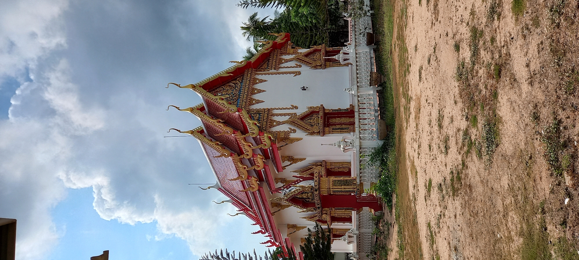 The image shows a Buddhist temple located in Moo 4, Thailand. The temple building is predominantly white with red and gold ornate detailing on the roof and exterior. The roof has multiple tiers with decorative curved spires at the edges. A cloudy sky is visible in the background. To the side of the temple, there's a grassy area with some trees and an expanse of dry, sandy ground. The photo appears to have been taken during the day.