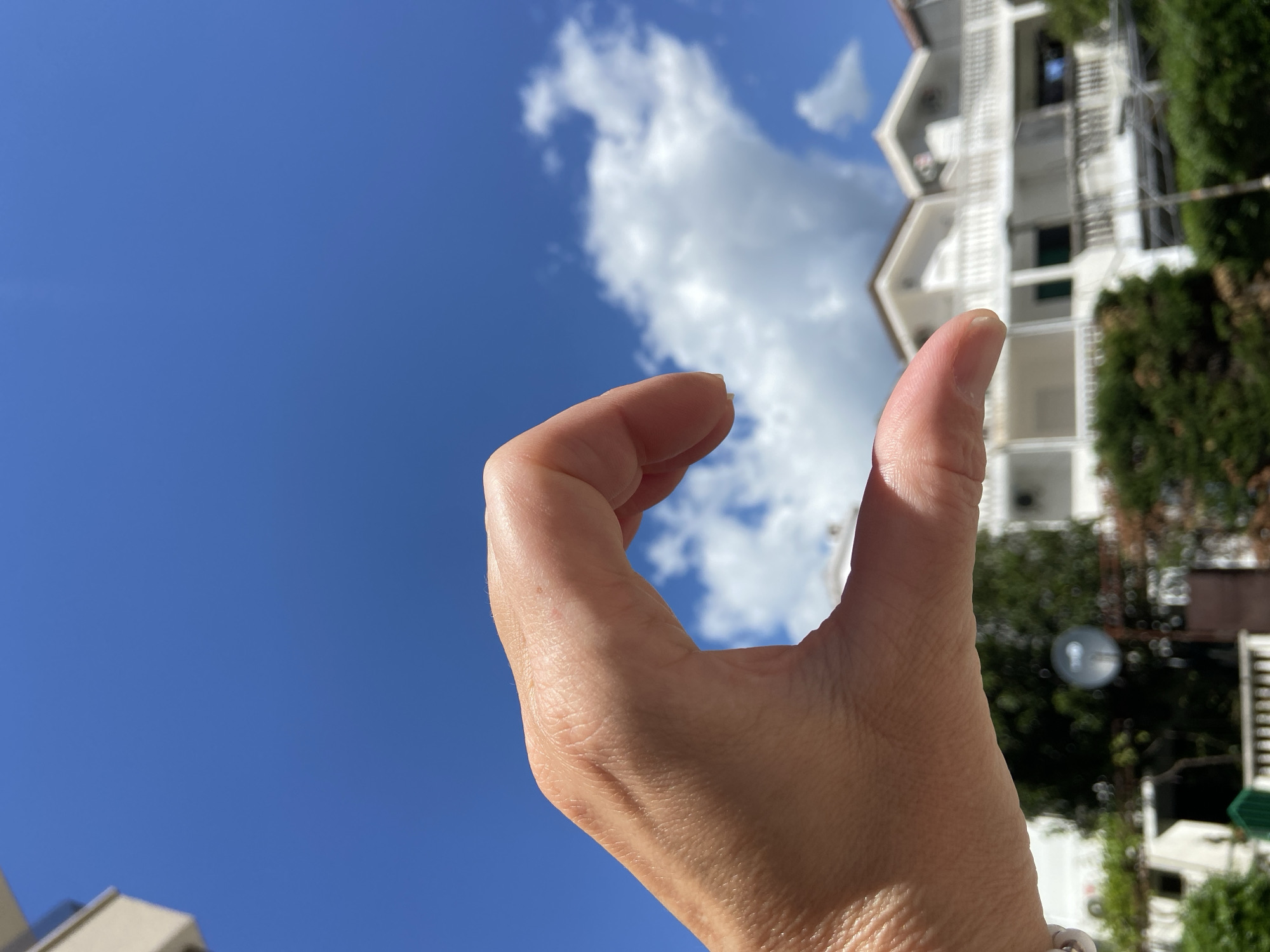 A hand is held up to the sky, with the thumb and forefinger forming an "OK" sign. The sky is bright blue with some white clouds. In the background, there is a white building with several balconies and green trees. The building appears to be located in the city of Budva, Montenegro.