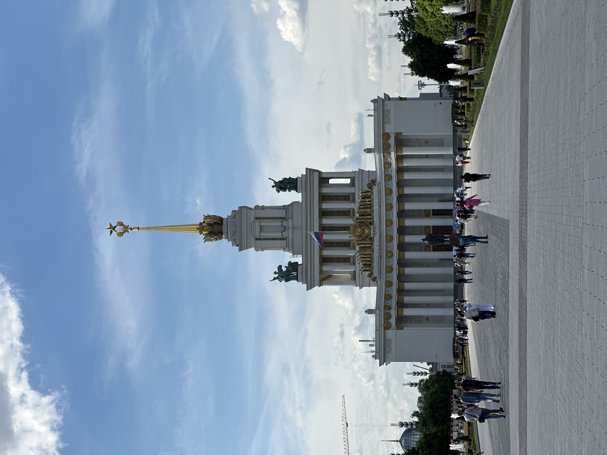 The image, originally rotated 90 degrees clockwise, captures a bright daytime scene at a grand architectural complex. The main subject is a large, ornate building, likely a pavilion, distinguished by its white facade and extensive golden architectural details. A prominent central spire, crowned with a golden five-pointed star within a wreath, rises from the building. The structure features multiple levels, decorative columns, and statues adorning its roofline, reflecting a grandiose architectural style. A small Russian flag is visible on the building's facade.
In the foreground, a wide, light grey paved plaza or walkway is occupied by several groups of people, who appear to be visitors or tourists enjoying a leisurely stroll. To the right of the paved area, a green landscape with trees, bushes, and active water fountains adds to the scenic appeal. The clear blue sky overhead is dotted with scattered white clouds, indicating sunny weather. In the background, other distant structures and a construction crane are visible, suggesting an expansive urban or exhibition complex.
Indistinct Cyrillic text is visible on the golden frieze of the building, appearing to read "НАРОДОВ СОЮЗ СОВЕТСКИХ СОЦИАЛИСТИЧЕСКИХ РЕСПУБЛИК" (Union of Soviet Socialist Republics). These architectural and textual details, combined with the overall grandeur, strongly suggest the location is the Exhibition of Achievements of National Economy (VDNKh) in Moscow, Russia. The scene conveys a calm and relaxed atmosphere, with individuals engaged in sightseeing and exploration.