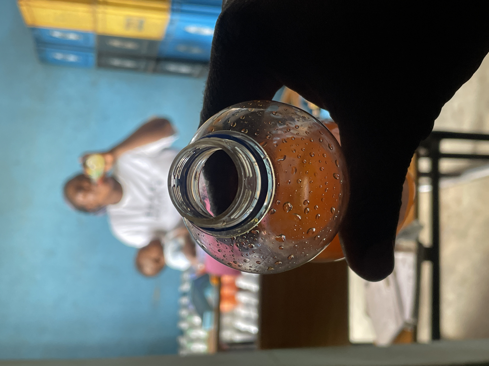 The image, originally oriented sideways, presents a close-up, low-angle shot, focusing on a hand holding a bottle while looking towards a blurred background. In the immediate foreground, a dark-skinned hand firmly grips a clear, cylindrical bottle filled with an amber-colored liquid, likely a beverage. The bottle's exterior is densely covered in condensation droplets, indicating it is a cold drink. The bottle's opening, with a distinct blue and white rim, is visible at the top center of the frame.
The background is significantly out of focus. To the left, a bright blue wall serves as a backdrop against which two blurry human figures are discernible. One figure appears to be an adult wearing a white t-shirt, holding an object in their hand. Below and slightly behind, a smaller, indistinct figure, possibly a child, is also visible. Above these figures, stacked colorful crates – yellow, blue, and black/dark grey – are arranged, commonly used for beverage transport and storage. Further to the right, faint outlines suggest shelving or racks.
The scene depicts someone holding a refreshing drink, possibly just purchased or about to be consumed. The setting appears to be an indoor commercial space, such as a small shop, kiosk, or a beverage distribution point, consistent with typical establishments found in Jos, Nigeria. The condensation on the bottle suggests warm ambient temperatures or recent refrigeration. The lighting indicates it is daytime. No discernible text is visible on the bottle or in the background due to the photographic blur.