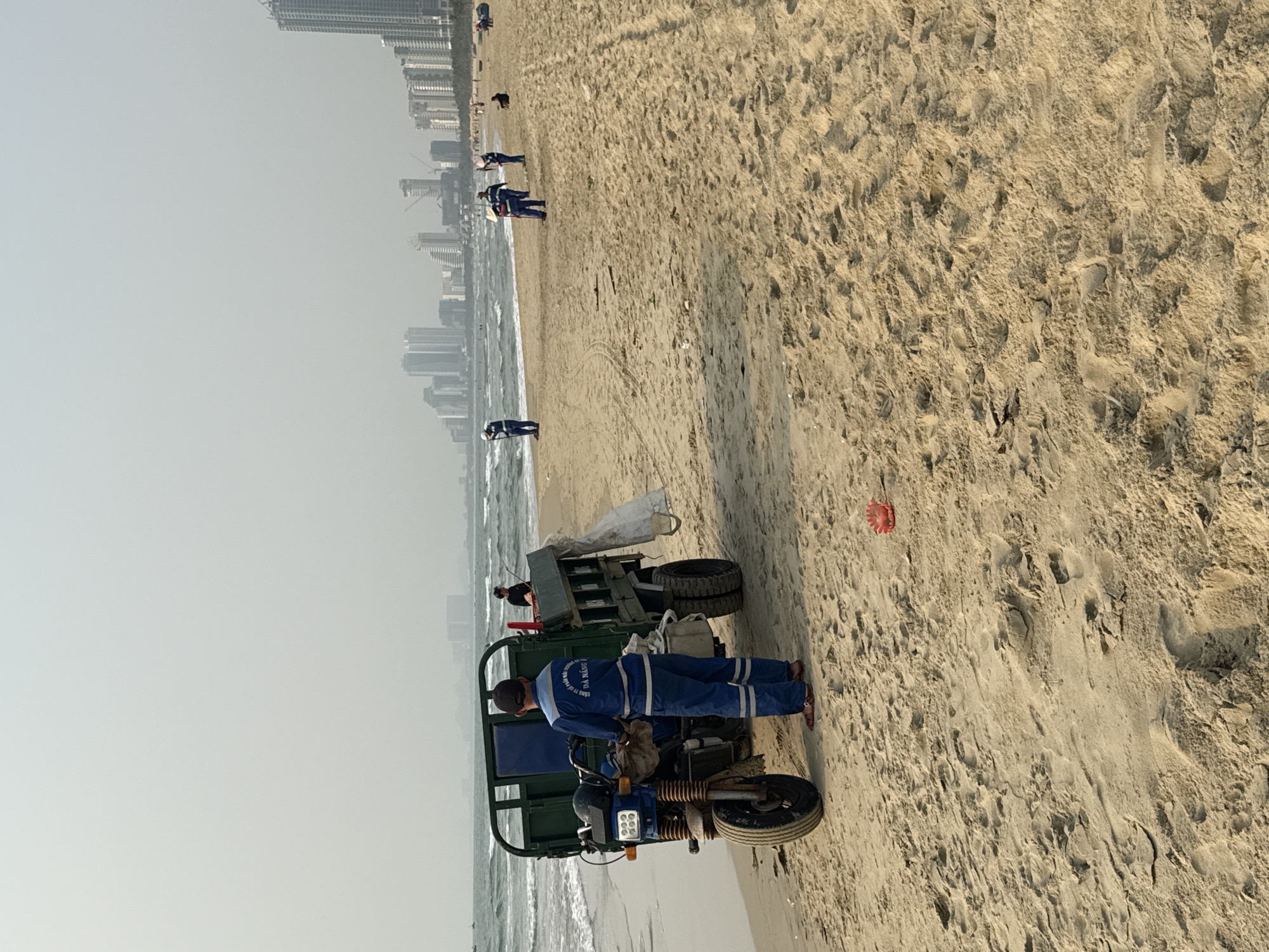 The image depicts a beach scene in Đà Nẵng, Vietnam. A worker wearing a blue uniform stands near a motorized tricycle with a trailer attached. The tricycle appears to be a utility vehicle used for cleaning or maintenance on the beach. The beach is sandy, with numerous footprints and tire tracks. In the background, there are several individuals walking along the shoreline. Further in the background, a city skyline is visible, indicating the urban setting of the beach. The sky is overcast and hazy, suggesting a somewhat humid atmosphere. The ocean water is lapping against the shore, with small waves. A possible piece of trash, red in color, can be seen in the sand.
