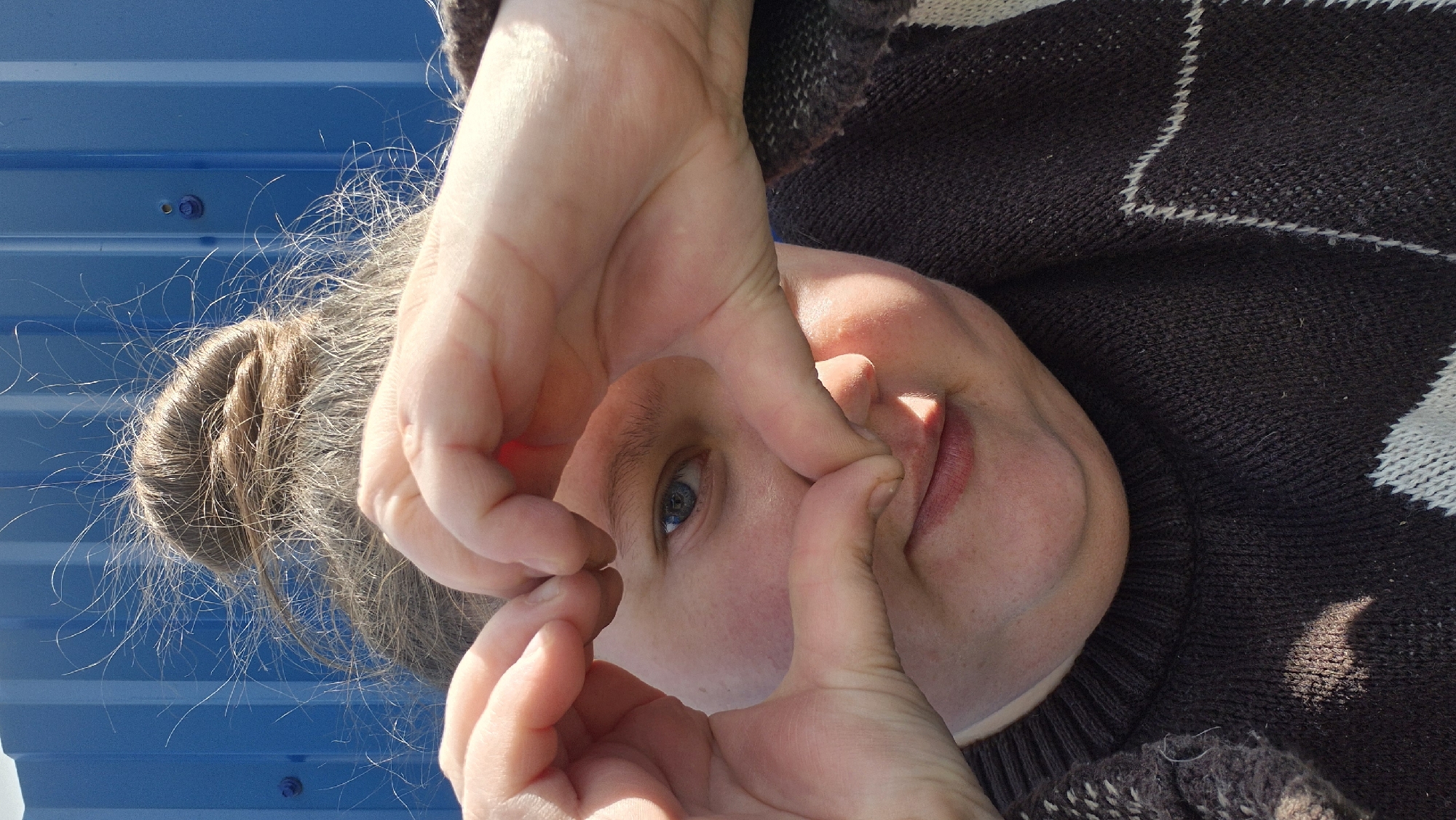 A middle-aged person is photographed from a low angle, with their face oriented towards the viewer and hands creating a circle framing their eye. They have fair skin, light blue eyes, and a closed mouth with visible pink lips. Their hair is pulled back in a bun, with stray hairs visible against a blue corrugated metal background. They wear a dark brown and white patterned sweater. The location is unknown.