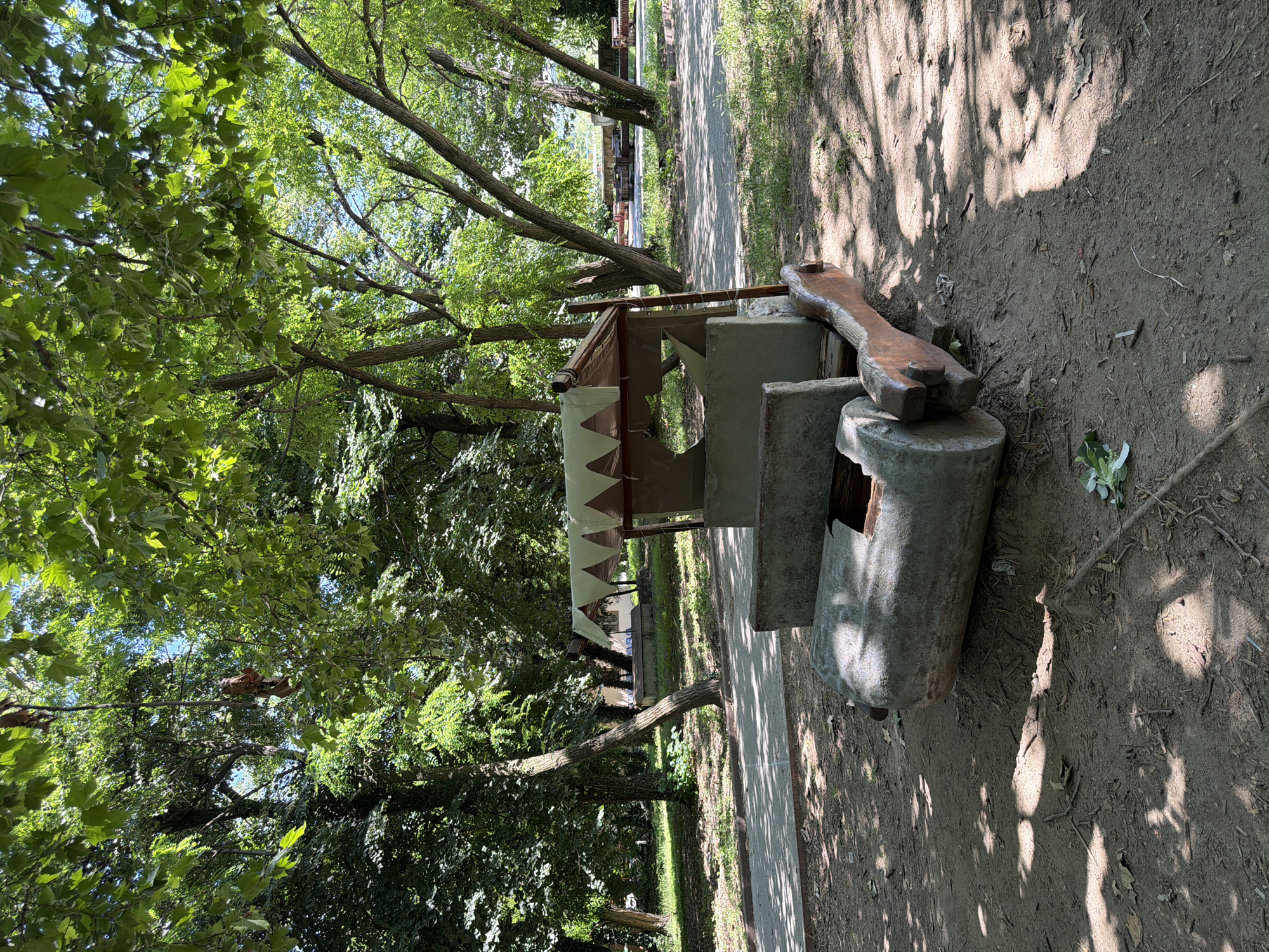 This is a vertical, eye-level shot of a makeshift playground attraction resembling a car, situated in a park in Pazardzhik, Bulgaria. The attraction is constructed from rough, unpainted concrete and wood, designed to evoke a prehistoric vehicle. The "wheels" are large, rough concrete cylinders, and the body appears to be made of concrete slabs and wooden planks. A tattered, triangular-patterned canvas canopy shades the top, supported by wooden poles. The attraction is placed on a dirt and leaf-covered ground, with a paved pathway visible behind it. The park setting is characterized by mature trees with lush green foliage, casting dappled shadows on the ground. In the background, other park amenities like benches and structures are visible, hinting at a public recreational area. No people are present in the image.
