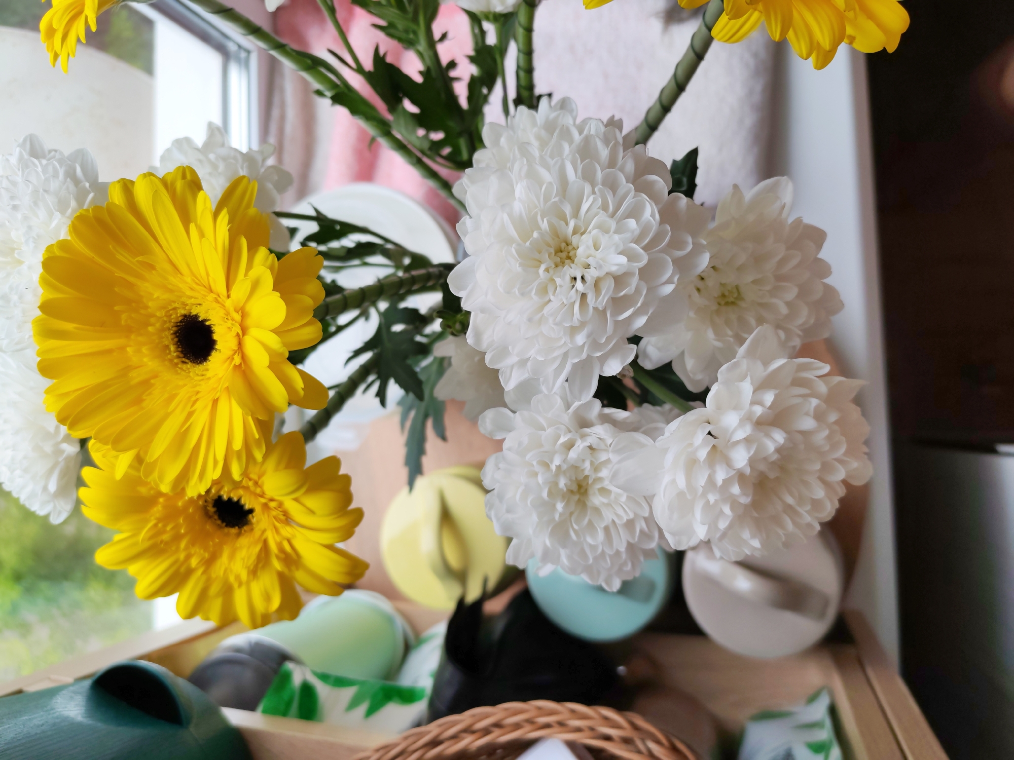 A close-up shot features a bouquet of yellow and white chrysanthemums in a basket. The yellow flowers, which appear to be gerbera daisies, are vibrant and have dark centers. The white chrysanthemums are full and fluffy, with many delicate petals. The bouquet is arranged with green stems and leaves.
The flowers are positioned in front of a window, with glimpses of greenery and a blurry background suggesting an outdoor scene. The light from the window illuminates the flowers, creating soft shadows and highlighting their textures.
Below the flowers, various objects are visible within the basket. These include colorful containers or lids in shades of green, blue, and yellow, as well as a black object that could be a small figurine or accent. A woven basket edge is prominent in the foreground, suggesting a rustic or natural setting. The overall impression is one of a bright and cheerful indoor arrangement.
The image is likely taken during the daytime due to the natural light filtering through the window. No people are visible in the frame. The setting appears to be a home or a decorative display. There is no visible text.
