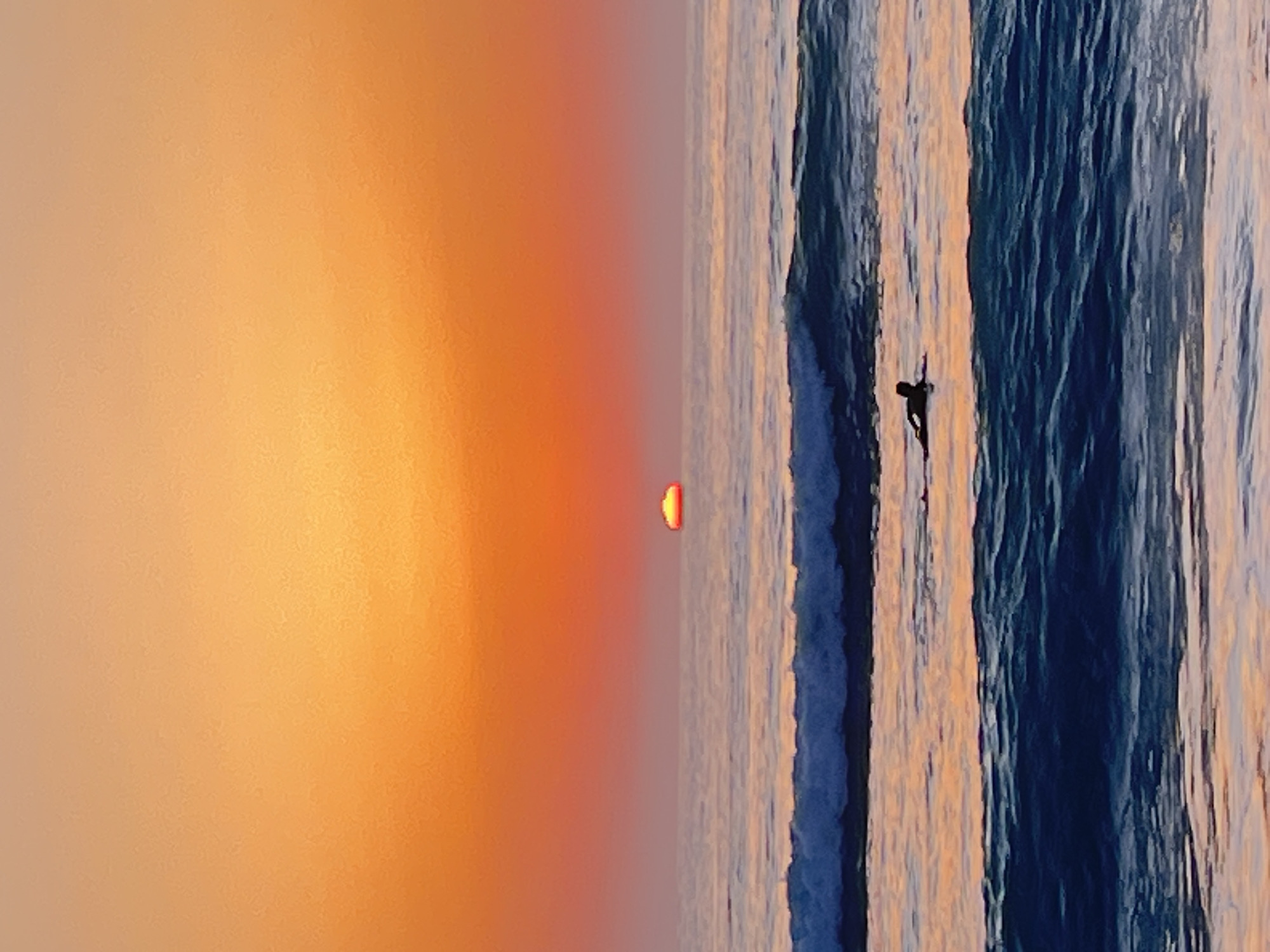 The image depicts a surfer riding a wave in the ocean, silhouetted against a sunset. The background features an orange sky transitioning into a gradient of colors closer to the horizon. The ocean appears textured with defined wave patterns, reflecting the colors of the sunset. A small, indistinct silhouette of a surfer is visible on the wave. The location is likely A Coruña, Spain, inferred from the sunset over the ocean and the presence of surfers, a common sight on the city's coast. The orientation of the image is vertical.